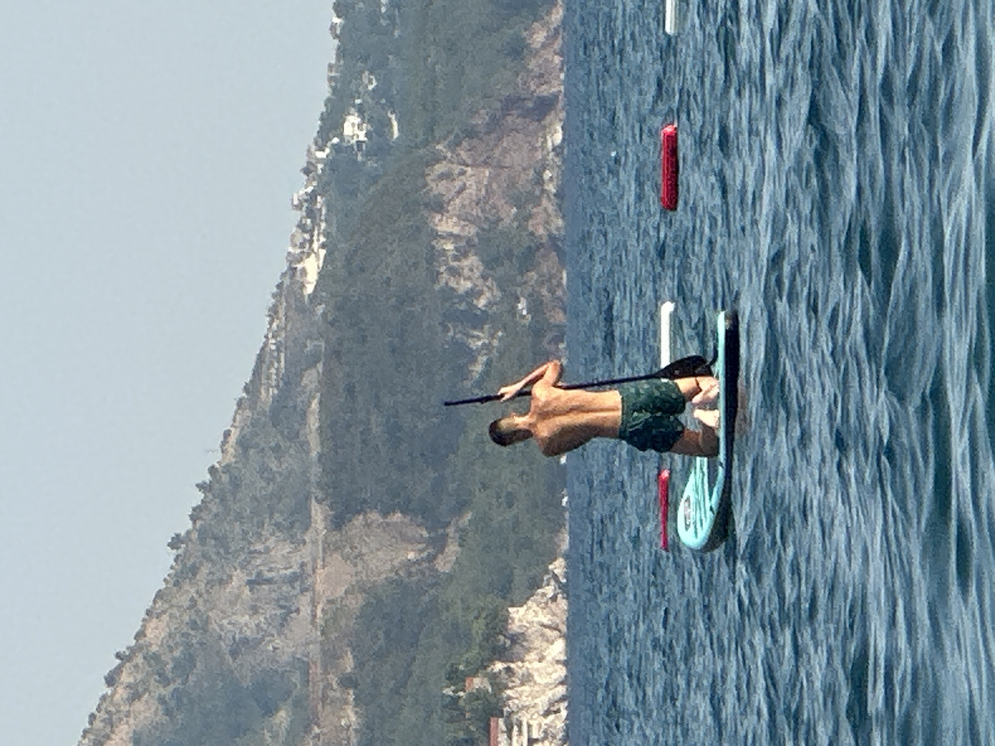 A man is paddleboarding in the sea off the coast of Boreti, Montenegro. He is on his knees on a paddleboard, holding a paddle. The water is blue and choppy, and there are hills in the background covered in trees and buildings. In the distance, there are also some red and white buoys in the water. The sky is clear and blue.
