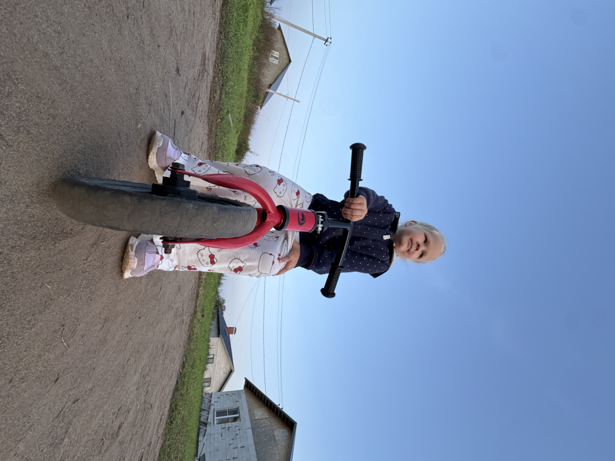 A young blonde girl wearing a dark blue hooded jacket with white polka dots and white pajamas with a Hello Kitty pattern sits on a red balance bike. Her feet are clad in light purple sneakers. She is looking up and to her right, with her hands on the handlebars. The balance bike is positioned on a dirt road. In the background, there is a grassy embankment with a few houses and utility poles. The sky is clear and blue, suggesting it is daytime. The setting appears to be a rural area, possibly on the outskirts of a village or small town, with the construction of new houses evident. The overall scene captures a child enjoying an outdoor activity.