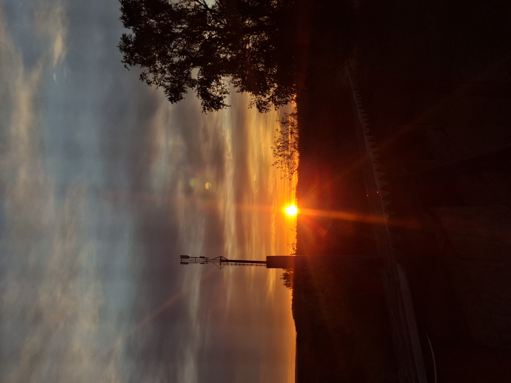 The image depicts a sunset with orange, yellow, and gray hues in the sky. A communication tower stands silhouetted against the setting sun. Trees and a road are visible in the foreground and on the horizon. The location is Unknown, Unknown.