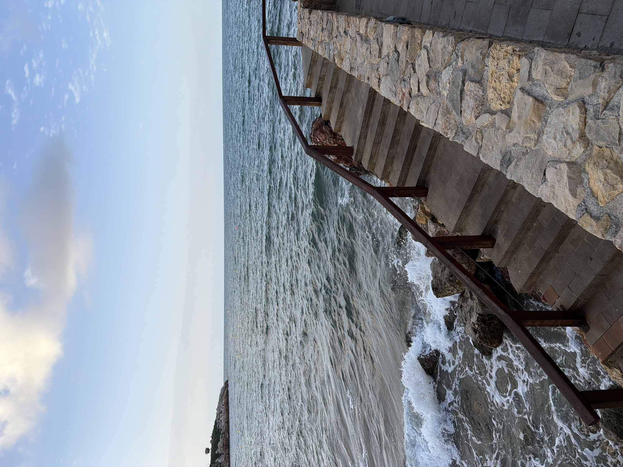 The image depicts an outdoor scene with a body of water and a stone-clad walkway. In the foreground, a set of concrete stairs with wooden railings descends towards the water, which is breaking against a rocky shoreline. To the right of the stairs is a low wall constructed from natural stones. The sea stretches to the horizon under a sky with scattered clouds. In the distance, a rocky outcrop with a lone tree is visible on the left. Yellow buoys are scattered in the water. This scene is located along the coastline of Salou, Spain, likely part of its promenade or coastal path system. No people are present.