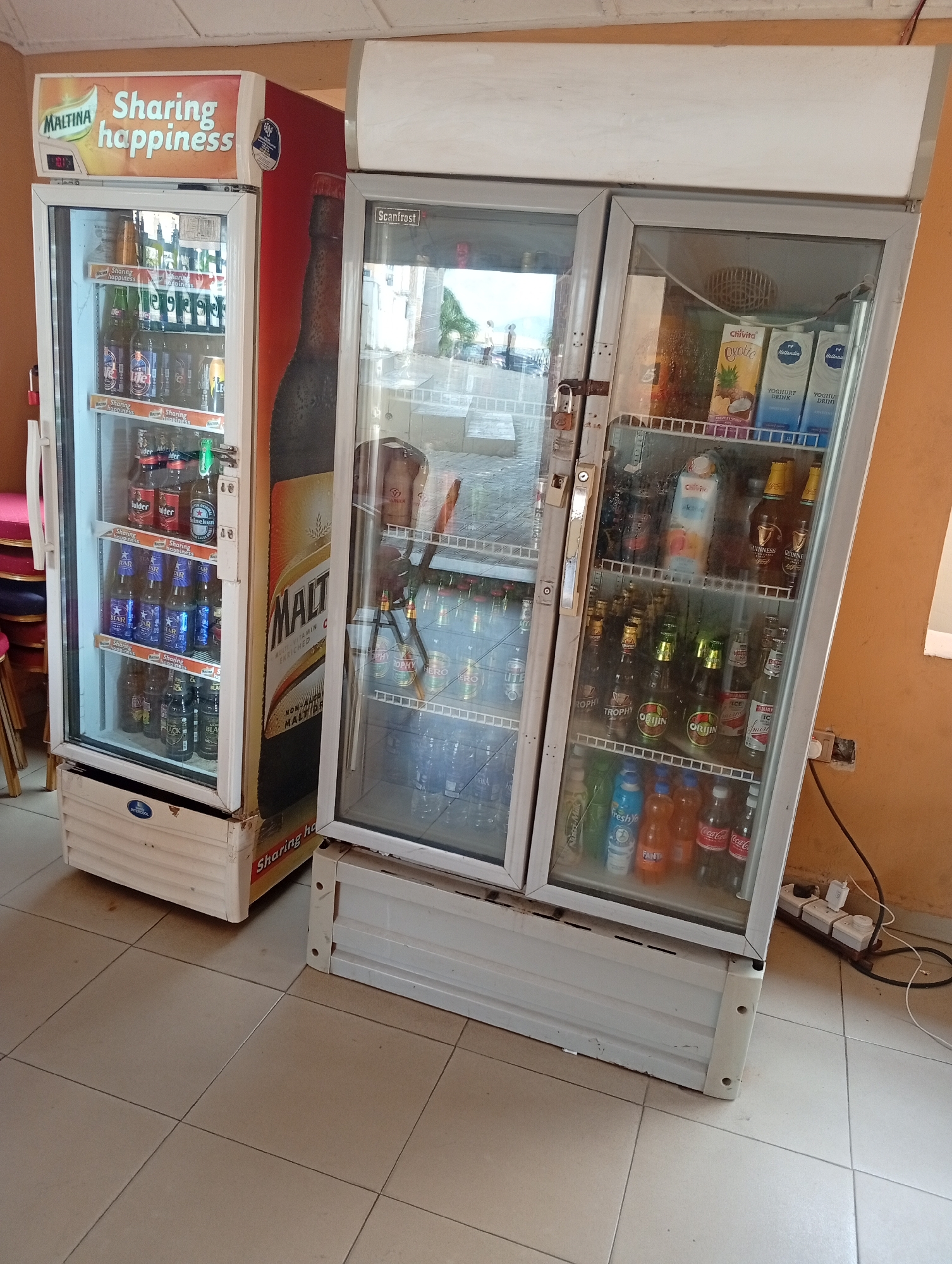Two refrigerated drink display cases stand side-by-side on a tiled floor. The display case on the left has "MALTINA Sharing happiness" printed on its top and side, showcasing various brands of bottled drinks inside, including "Gulder," "Heineken," "STAR," and "Black." The display case on the right has "Scanfrost" at the top and contains brands like "Chivita," "Hollandia," "Guinness," "Trophy," "Orijin," "Smirnoff Ice," "Fresh Yo," "Fanta," and "Coca-Cola." An electrical outlet strip is near the base of the right display case. The scene is likely in Lokoja, Nigeria, given the local beverage brands displayed.