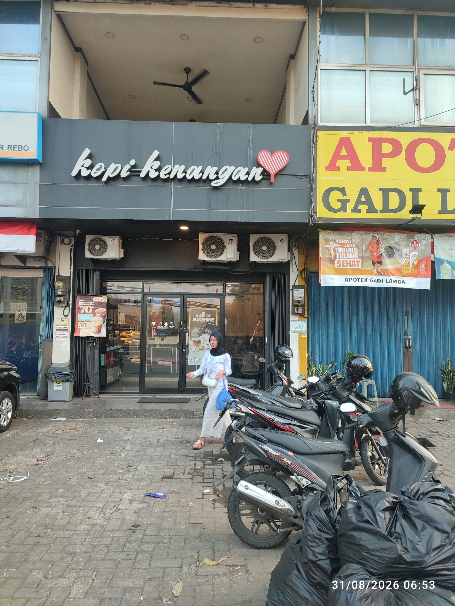 The image shows the exterior of a "Kopi Kenangan" coffee shop in East Jakarta, Indonesia. A woman in a light-colored hijab and white clothing walks away from the entrance, carrying a white purse and a blue bag. In front of the shop, several motorcycles are parked, some covered with helmets. Large black trash bags are piled on the sidewalk to the right of the motorcycles. A banner for "Apotek Gadi Lamba" (Gadi Lamba Pharmacy) is visible above the coffee shop. The street in front of the establishment is paved with gray brick. The timestamp "31/08/2026 06:53" is present in the lower right corner.