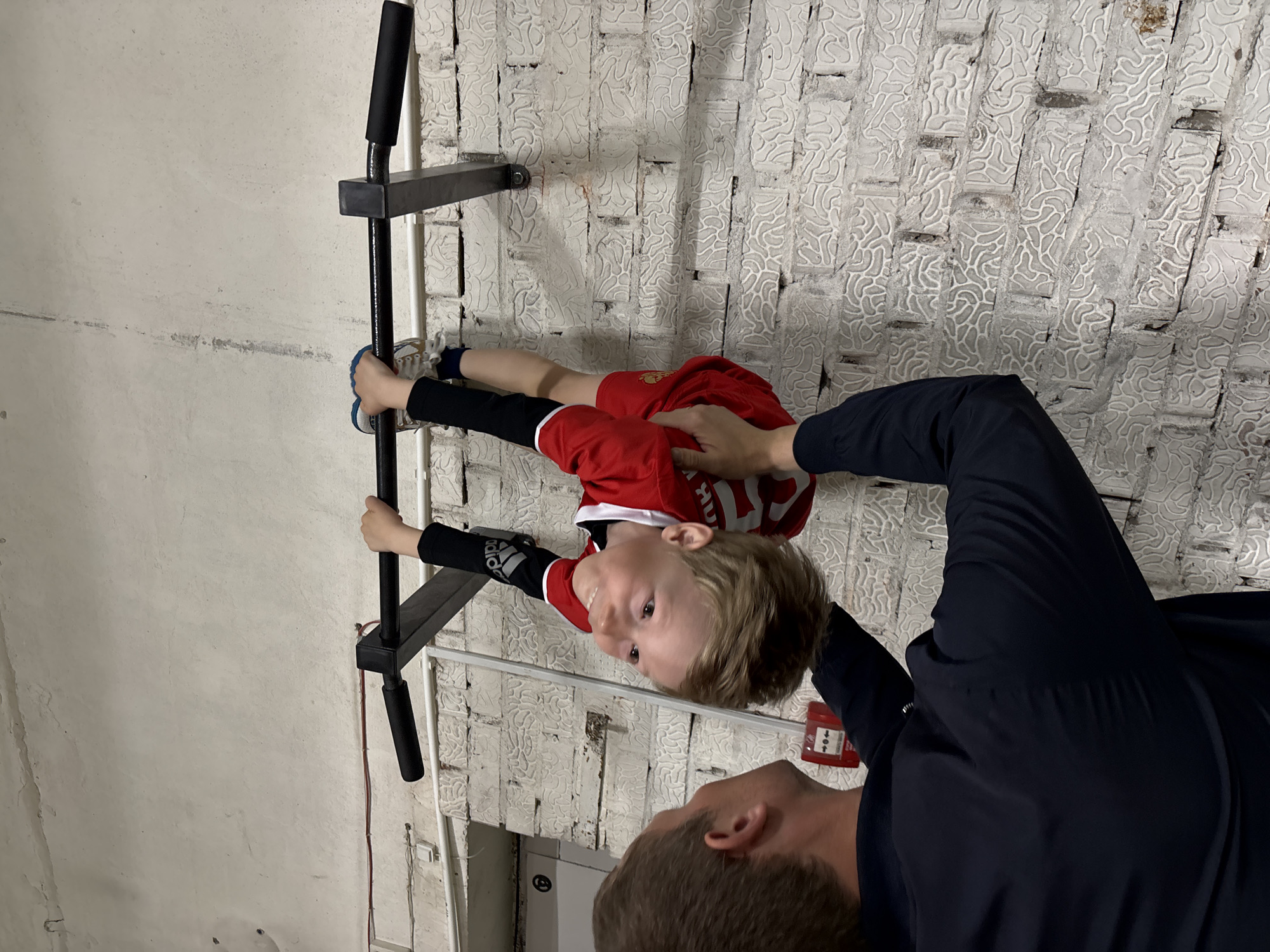 A small boy in a red shirt and black leggings hangs from a black pull-up bar attached to a white, textured wall. He is being supported by an adult who is partially visible, wearing a black shirt. The location appears to be indoors, with a white wall that may be a basement. A red fire alarm box is visible. The boy's feet are positioned toward the plain white portion of the wall, while the adult is mostly obscured by the right-hand side of the frame. The photo has been rotated 90 degrees clockwise. The location is given as the city of Chaikovsky, Russia.
