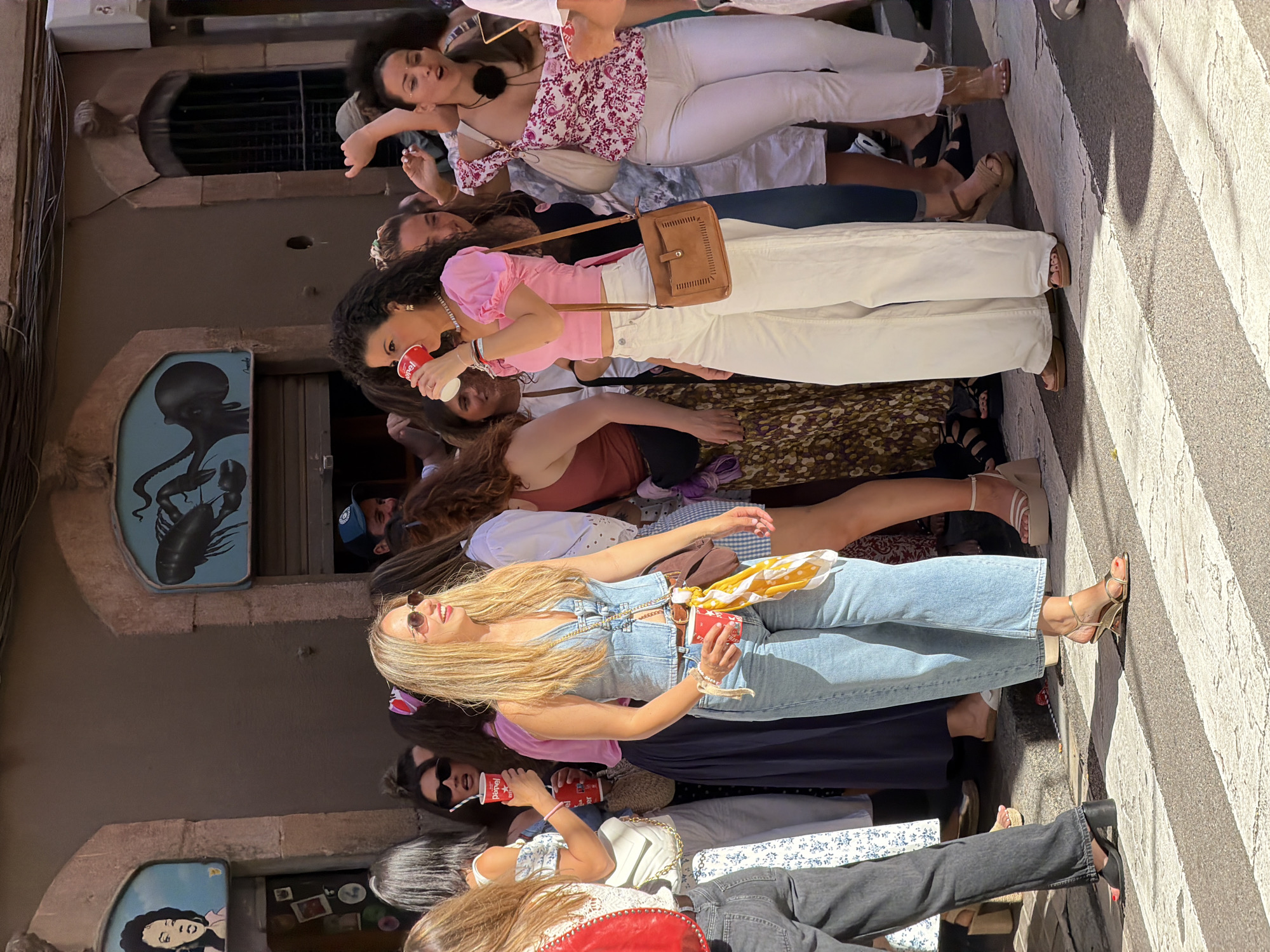The image depicts a group of young women in what appears to be a busy street in Barcelona, Spain, possibly on Las Ramblas, a popular pedestrian street. The women are dressed in casual, summery attire, suggesting a festive or celebratory occasion. Two women in the foreground are holding red cups and seem to be engaged in conversation or enjoying a drink. One woman on the left is wearing a denim vest over a white top and light blue jeans. The other woman on the right is wearing a pink puff-sleeve top and wide-leg white pants. In the background, other individuals are visible, some holding drinks and looking towards the camera. The street scene includes a building with a sign featuring a caricature of a woman and another with a depiction of a lobster. The ground is paved with white and gray stripes, characteristic of a pedestrian crossing. The lighting suggests it is daytime with bright sunlight casting shadows.