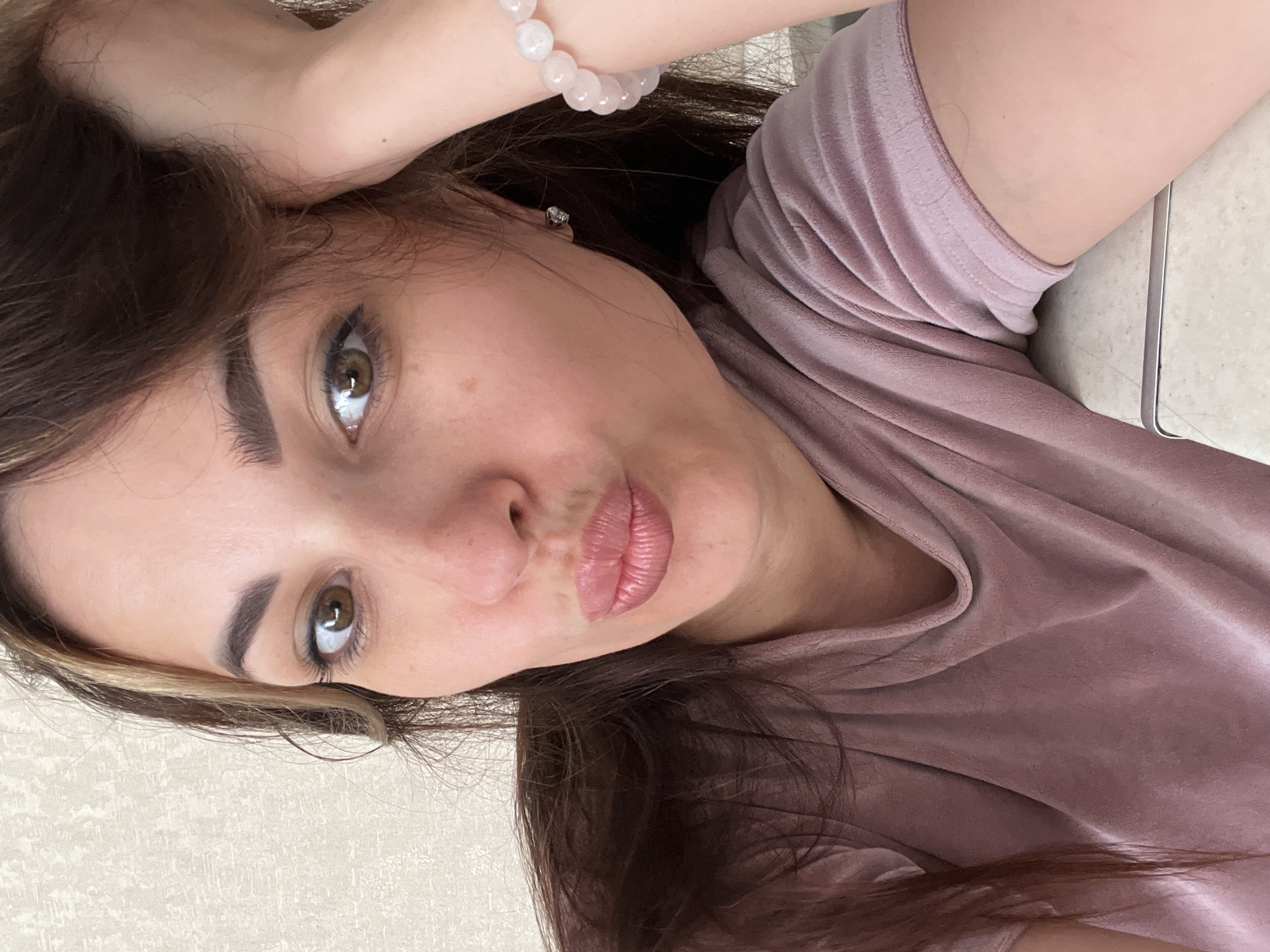 A woman with brown hair and brown eyes is posing for a selfie. She is wearing a pink t-shirt and a pink beaded bracelet on her left wrist. Her right arm is bent and resting on her head. Her lips are pursed in a pout. The background is a textured beige wall.