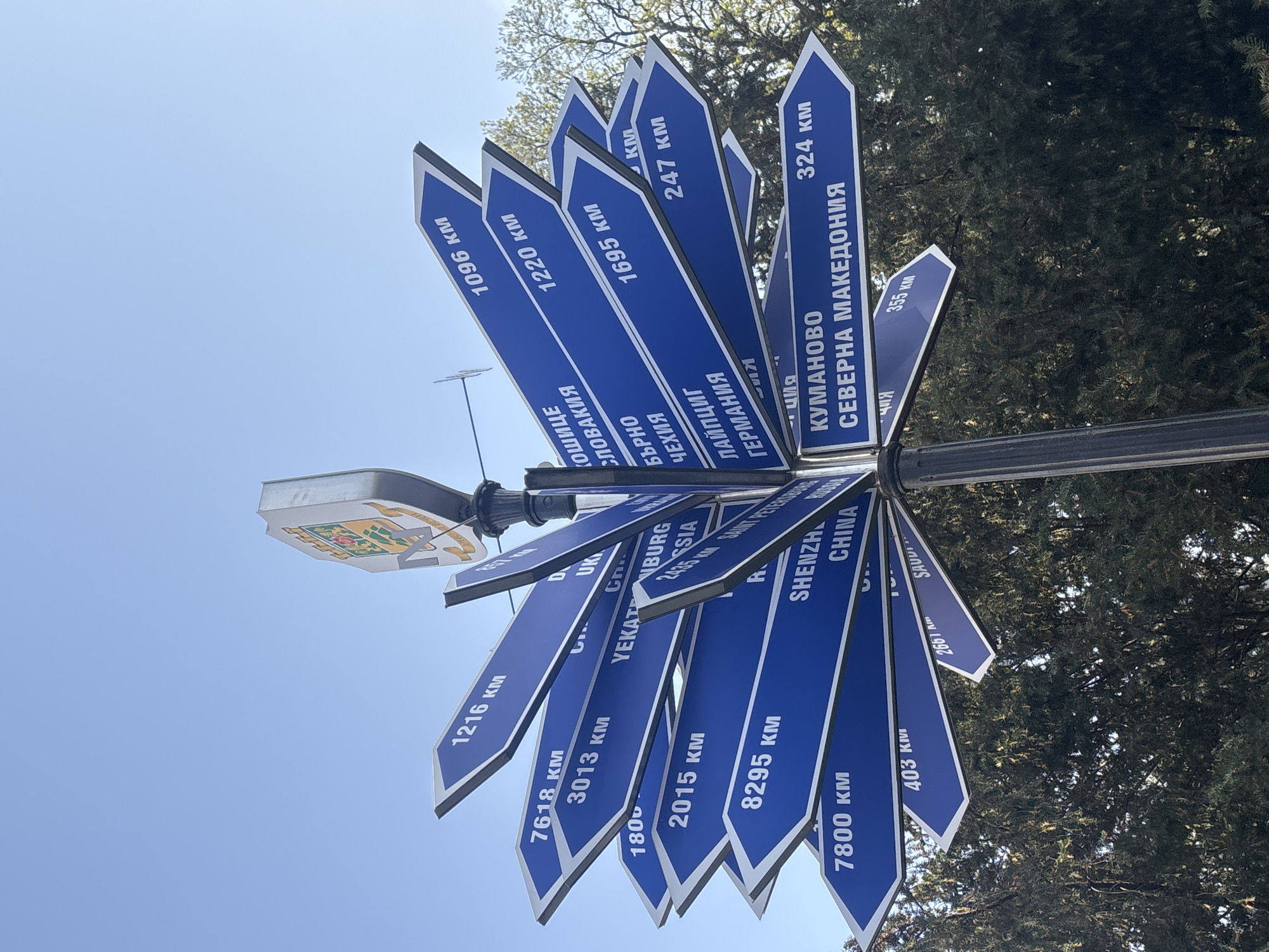The image shows a signpost with numerous blue signs pointing in different directions, each indicating a city and the distance in kilometers. Locations listed include Kosice, Slovakia; Brno, Czech Republic; Leipzig, Germany; Kumanovo, North Macedonia; Yekaterinburg, Russia; Saint Petersburg, Russia; Shenzhen, China; and Saudi Arabia. The signpost is located in Plovdiv, Bulgaria, as evidenced by the city's coat of arms at the top of the post. The sky is visible in the background, with trees surrounding the signpost.