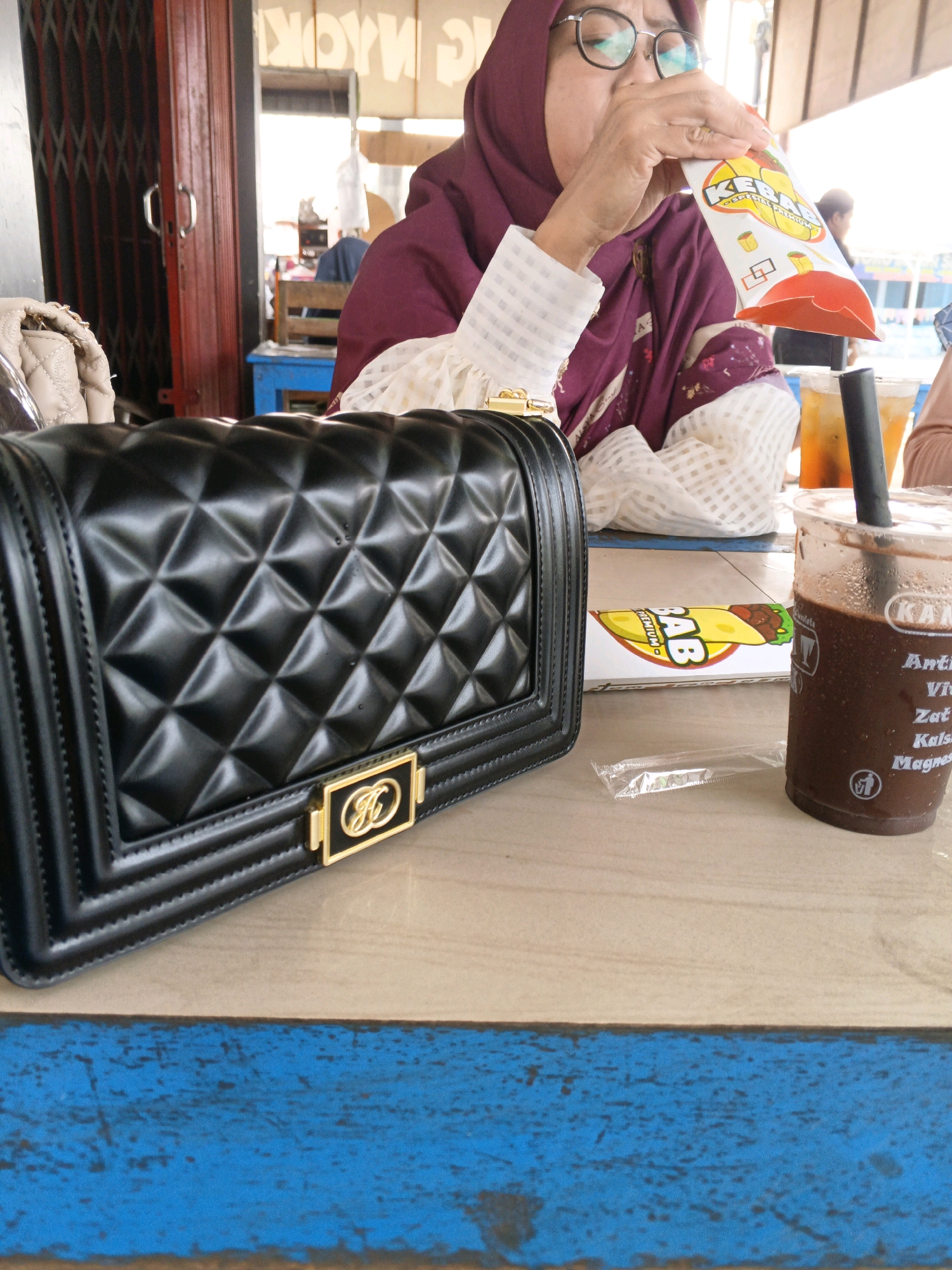 In Pungguk Lama, Indonesia, a woman wearing a hijab and glasses is seated at a table with a black quilted handbag and a beverage. The handbag is in the foreground, taking up the left side of the image. It has a quilted diamond pattern and a gold clasp. To the right of the handbag is a cup with a dark liquid and a straw, appearing to be a chocolate drink. Behind the cup and handbag, a box with "KABAB" written on it is visible. The woman is in the background, partially obscured, holding a small white and orange packet and drinking from it. She is wearing a burgundy hijab and a white top with intricate patterns. The table surface is made of wood with a blue painted edge. In the background, a red metal gate and blurry figures of people suggest a casual, outdoor or semi-outdoor dining area.