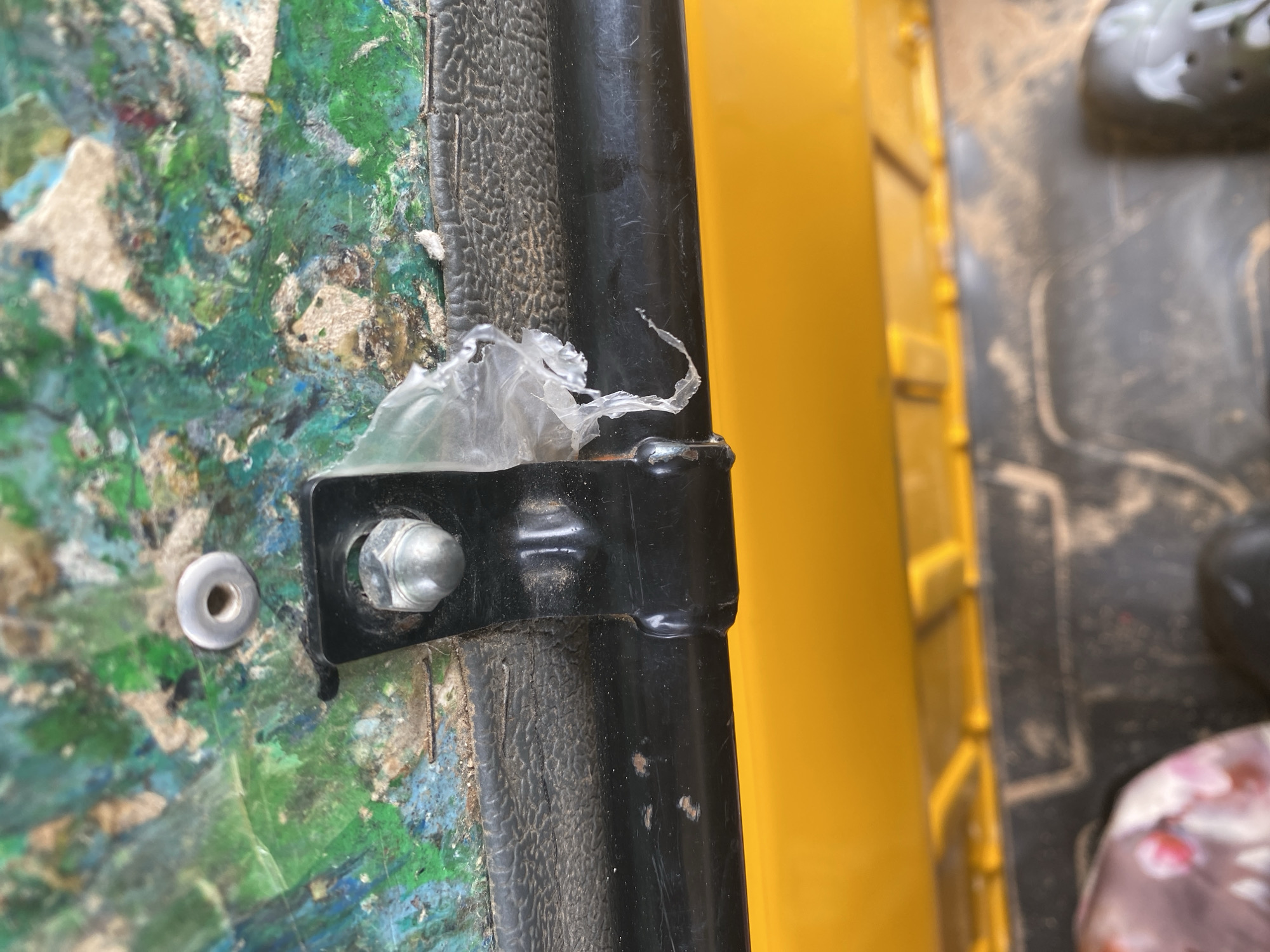 The image shows a close-up view of a part of what appears to be a vehicle or industrial equipment, possibly outdoors. The main subject is a black metal bracket attached with a bolt and nut to a textured, multi-colored surface. This surface is a rough composite material with patches of green, blue, and white paint or coating, suggesting wear and tear or a deliberate artistic design. There are also small white debris-like specks and a torn piece of translucent plastic film caught on the bracket.
To the right of the bracket, a thick black cylindrical rod or pipe is visible, partially obscuring a bright yellow surface in the background. This yellow surface has a ribbed texture, possibly indicating a plastic or metal component. The lighting suggests it is daytime, but no specific time of day can be determined. There are no people visible in the frame.
The overall impression is one of a functional, perhaps utilitarian, object that is exposed to the elements or heavy use, given the worn appearance of the painted surface and the presence of debris. The image is focused on the detail of the attachment and surrounding materials, offering no clear indication of a specific activity or event. The location context of Jalingo, Nigeria, does not provide any directly visible cues within the image itself, but suggests a potentially warm climate where such equipment might be used. No text is visible in the image.