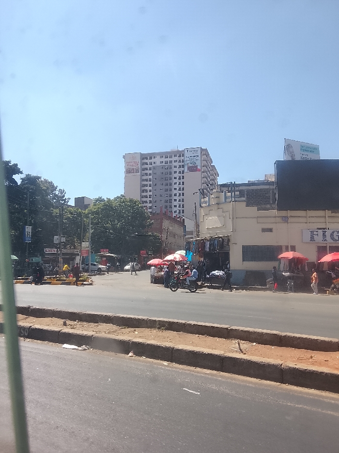 A bustling street scene under a clear sky features pedestrians and motorcycles near crosswalks and red parasols. A tall building displays "Park Inn" on its side, while a sign reading "FIG..." is visible on a lower structure. Street vendors are present, selling clothing. The foreground includes a curb separating the street from a dirt shoulder. The photo appears to be taken from a moving vehicle.