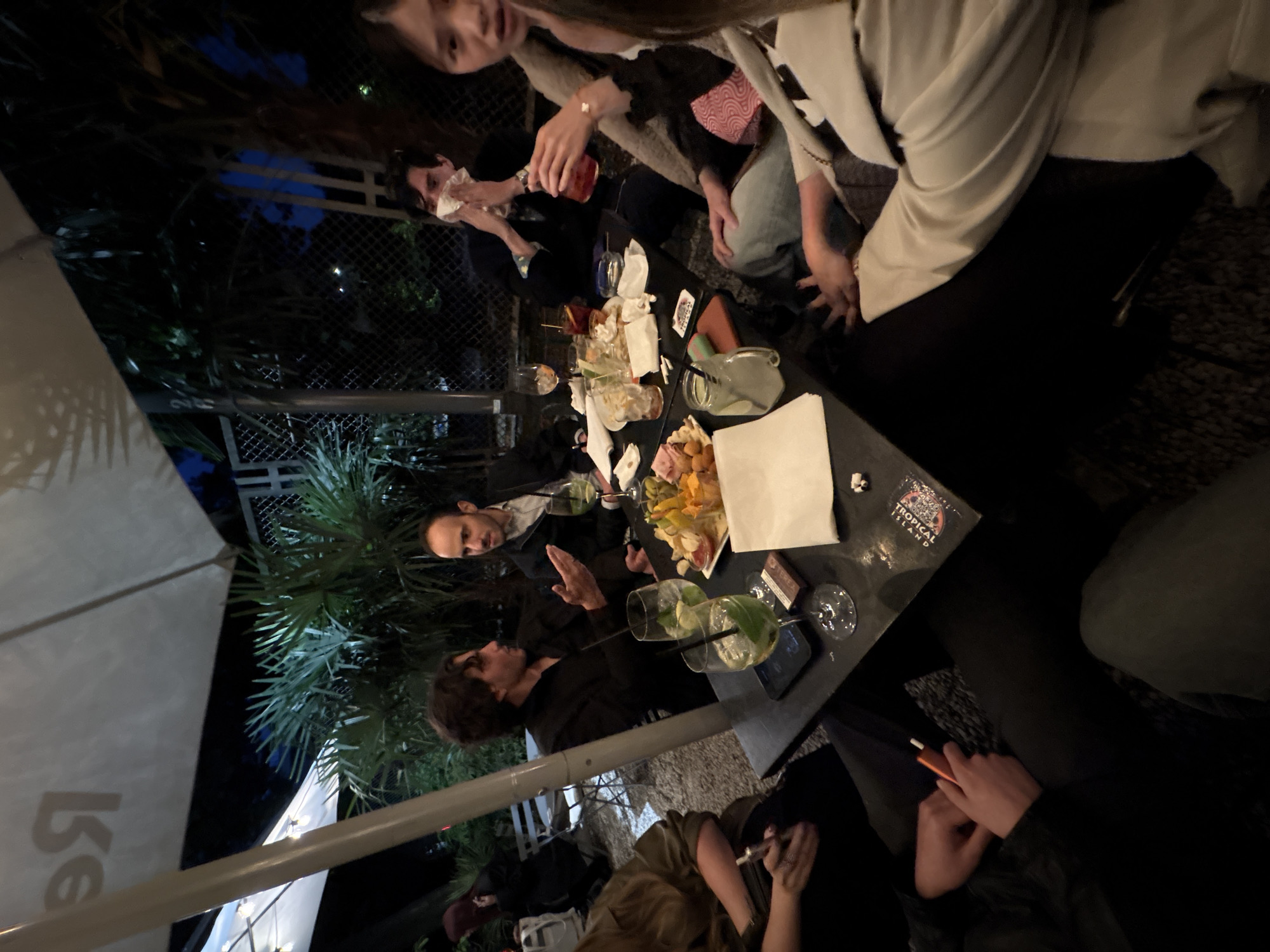 A group of people are gathered around a square table at an outdoor bar or restaurant, likely in Milan, Italy. The table is covered with glasses, some containing drinks with lime wedges and straws, a plate of food (possibly a charcuterie board or appetizer), napkins, and other miscellaneous items. There are at least eight people visible in the frame, some seated and some standing. Some are engaged in conversation and others are holding drinks or cigarettes. The setting is nighttime, illuminated by artificial lighting and partially shaded by a large umbrella. A metal fence and some foliage can be seen in the background.