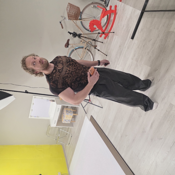 The image captures an indoor scene in what appears to be a photography studio located in Saint Petersburg, Russia. A male subject, estimated to be in his 30s-40s, with curly light brown hair and facial stubble, stands facing slightly right of the camera's perspective but looking directly at the viewer with a neutral expression. He is wearing a dark brown short-sleeved top with an intricate patterned texture, loose-fitting dark grey trousers, and white socks. His left hand holds an orange-cased smartphone, while his right hand holds a small, dark, unidentified object.
The environment features light grey walls, with a distinct bright yellow section on the far left. The floor is covered with light-colored, wood-grain laminate. On the left, a white photography backdrop sheet is partially unrolled on the floor. A tall black stand with a white softbox is positioned near the left wall. In the background, a white vanity desk with an illuminated mirror and a small, round stool with a yellow and grey pattern is visible. Further to the right in the background, two bicycles are present, one a cream or beige color with a wicker basket on its handlebars. A black camera tripod stands in front of the bicycles, and a red rocking horse is positioned on the floor between the tripod and the viewer. A dark, rectangular object, possibly part of a stand or reflector, is partially visible in the lower right foreground.