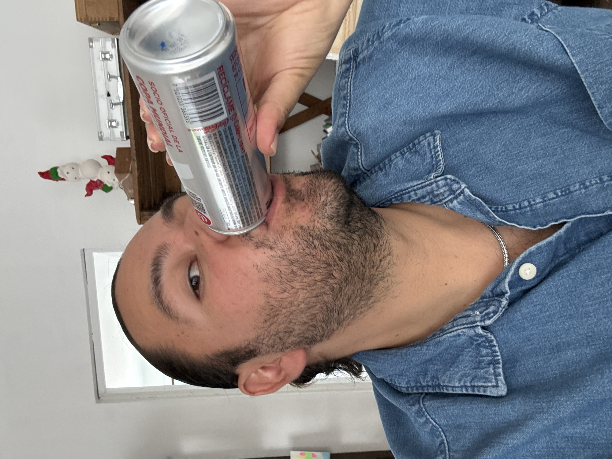 A person with dark hair and a beard is holding a can of Coke Light with Spanish text on the label, including "Socio Oficial de la Copa Mundial," indicating it is an official sponsor of the World Cup. The label also has "RECICLAME" and a barcode. The person is wearing a blue denim shirt. The environment includes a white wall with a window, a wooden shelf with Christmas decorations. The location, Puebla, Mexico, is inferred from the Spanish text on the Coke Light can.