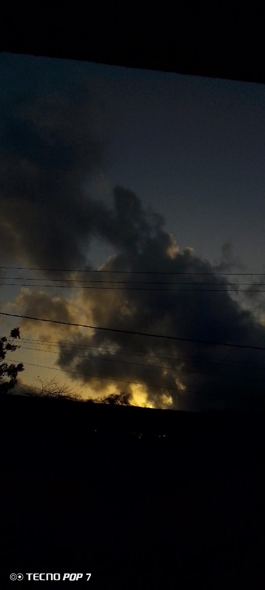The image shows a dark sky with dramatic, stormy clouds. A bright yellow glow is visible behind the clouds, suggesting the setting or rising sun. Several power lines stretch horizontally across the frame. In the foreground, silhouetted against the sky, is a dark, shadowy landscape that appears to be a hillside or rooftop, with sparse, bare tree branches visible on the left side. The bottom right corner of the image contains a watermark indicating "TECNO POP 7". The image was taken in Altagracia, Venezuela.