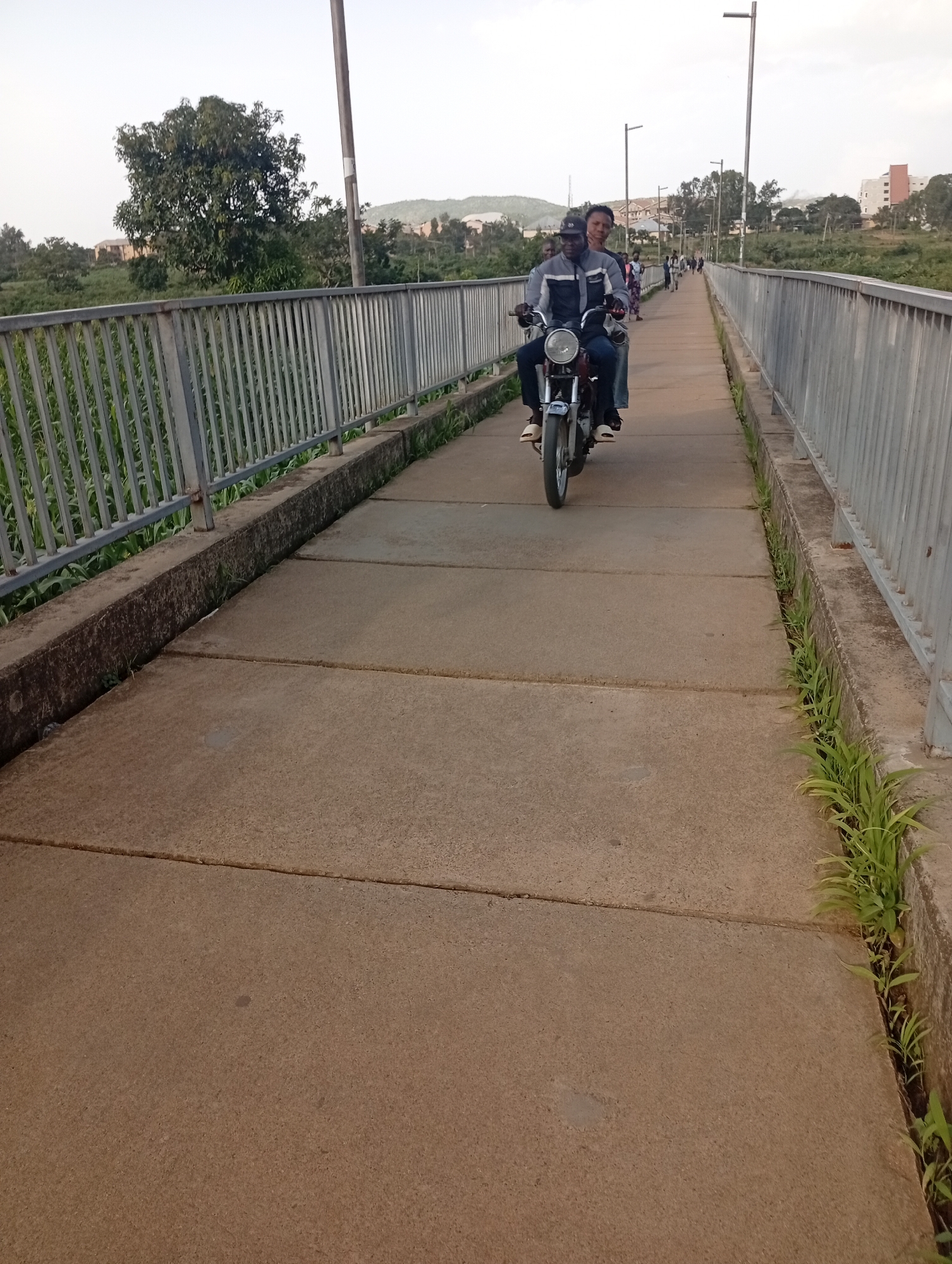 The image depicts a concrete walkway on a bridge in Rafin Par, Nigeria, with a metal railing on both sides. In the foreground, the concrete surface is textured with visible expansion joints. A man and a woman are riding on a motorcycle, with the man driving. They are moving away from the camera. The woman is sitting behind the driver, her left arm resting on his shoulder. Both are wearing casual clothing. Further down the bridge, several other pedestrians are visible in the distance, walking in the same direction. The background features lush green vegetation, trees, and some buildings, suggesting an urban or semi-urban environment. The sky is overcast.