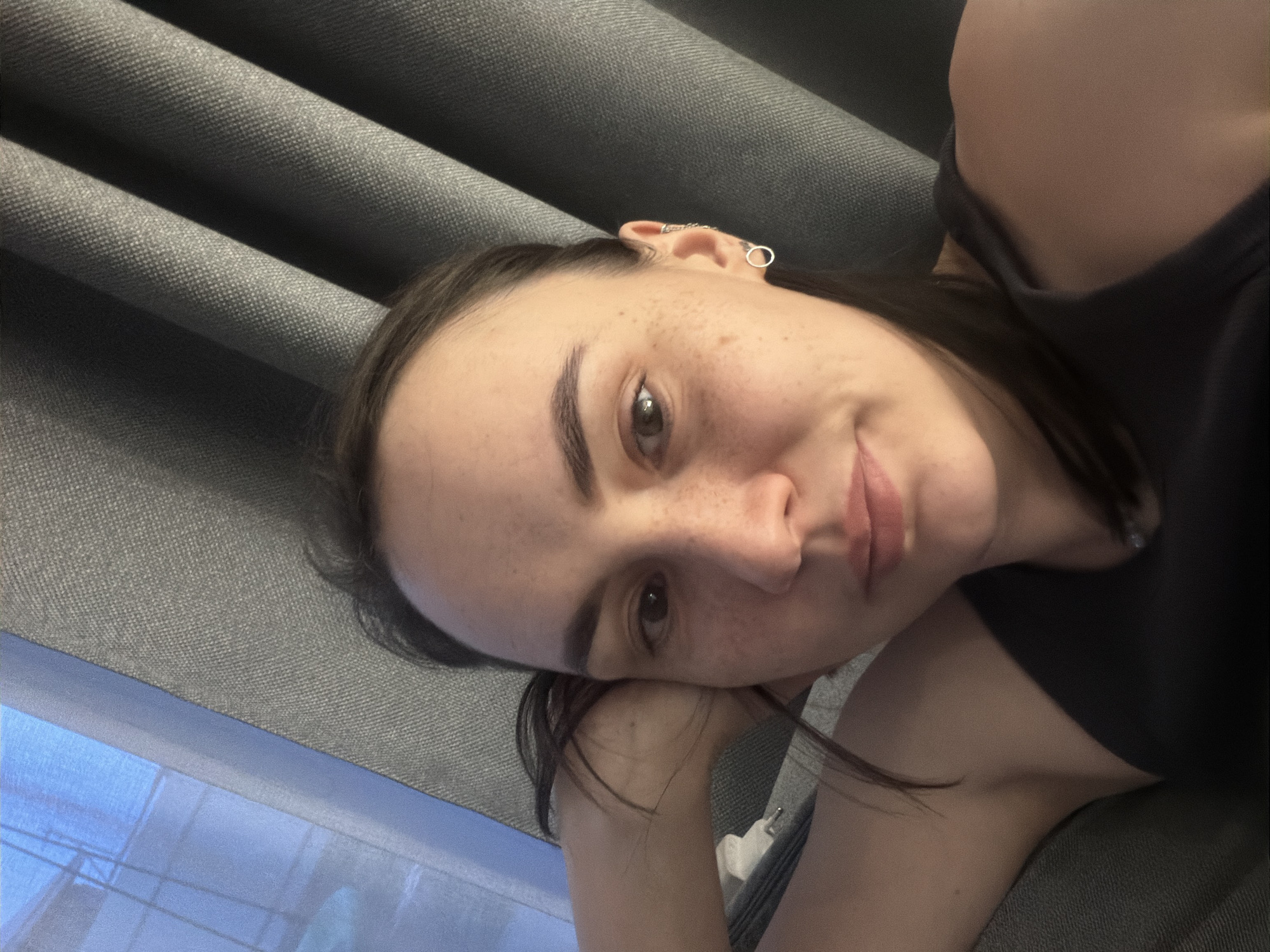A medium shot captures a woman lying down. She has dark hair, fair skin with freckles, and is wearing earrings. She is resting her head on her hand, which is visible in the lower-left corner. She is wearing a dark-colored top. The background consists of gray fabric, possibly curtains or upholstery, and a glimpse of a blue, reflective surface, potentially a window. There is no indication that this scene is specifically in Chaikovsky, Russia.
