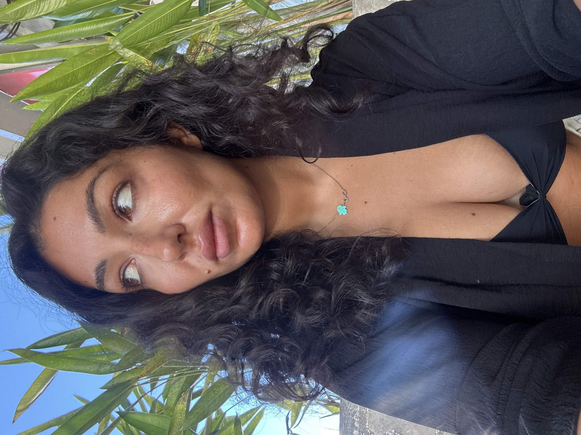 A medium shot shows a woman with dark curly hair wearing a black top with a knot on the chest and a black cardigan. She has a necklace with a blue charm. The background includes green leaves, a blue sky, and a concrete structure. The photo appears to be taken outdoors in a tropical or subtropical climate, possibly in the Kuta Selatan area of Indonesia, based on the presence of tropical foliage. The woman is looking slightly off-center with a thoughtful expression.