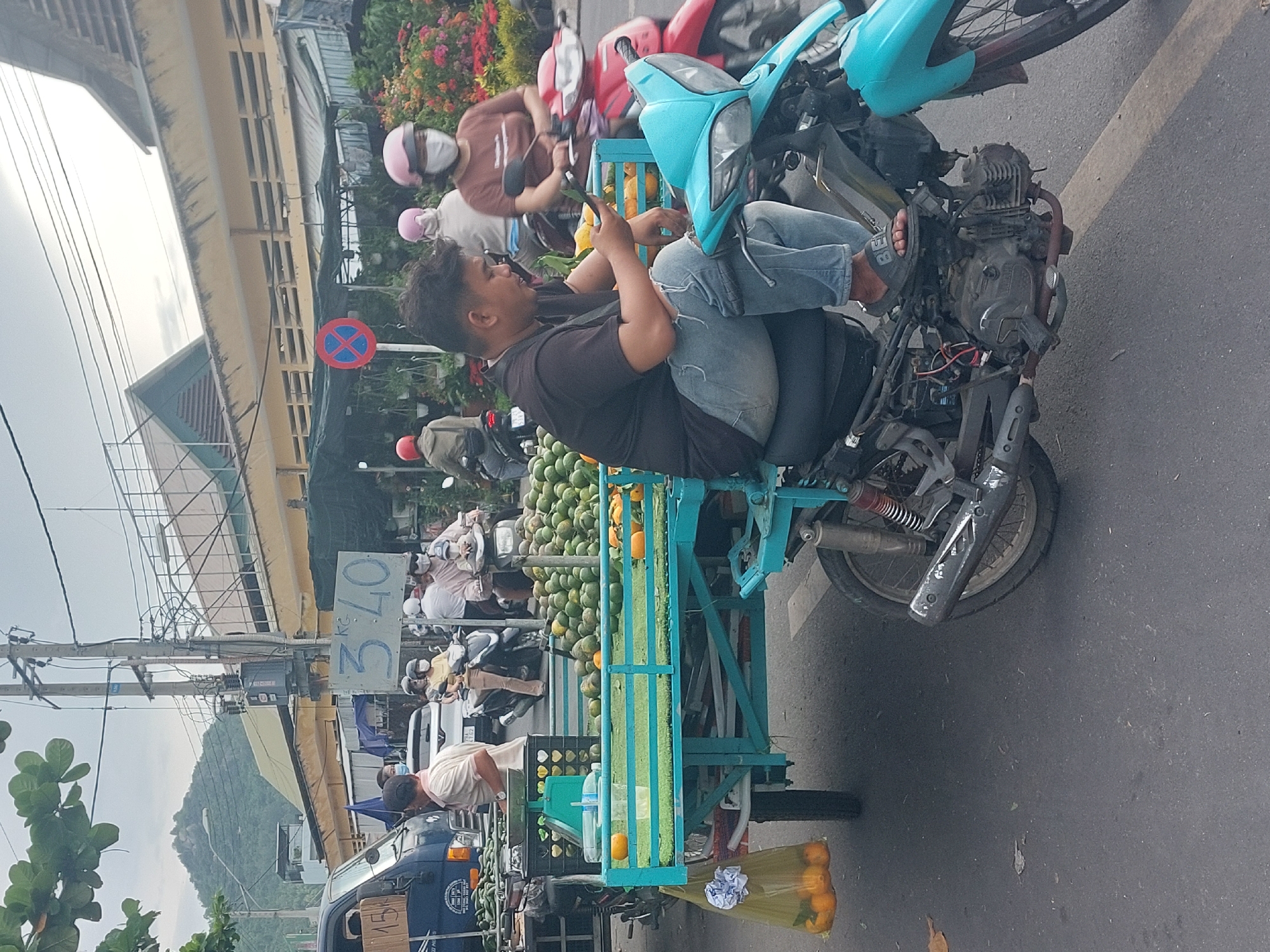 A man sits on a turquoise motorcycle, looking at his phone. The motorcycle is attached to a trailer filled with oranges and other fruits. The trailer is also carrying green artificial grass. In the background, there are other motorcycles, vendors, and buildings. The scene appears to be a street market in Nha Trang, Vietnam. The man is wearing a black t-shirt and ripped blue jeans, with a pair of black sandals. The trailer is blue and green. The fruits are mostly oranges. There are also some green, round fruits in the background. The buildings in the background are yellow and brown. The sky is overcast.