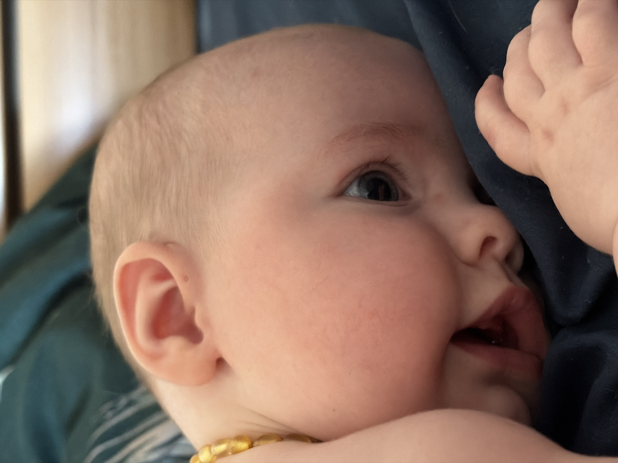 A close-up shot shows a baby lying on their side, seemingly indoors. The baby has fair skin, light hair, and is wearing what appears to be an amber necklace. Their mouth is slightly open, and their eyes are directed upwards. Part of the baby's hand is visible in the upper right corner, resting against a dark blue fabric. The background includes glimpses of a striped surface and what appears to be a dark green or blue cloth. Location information not available.