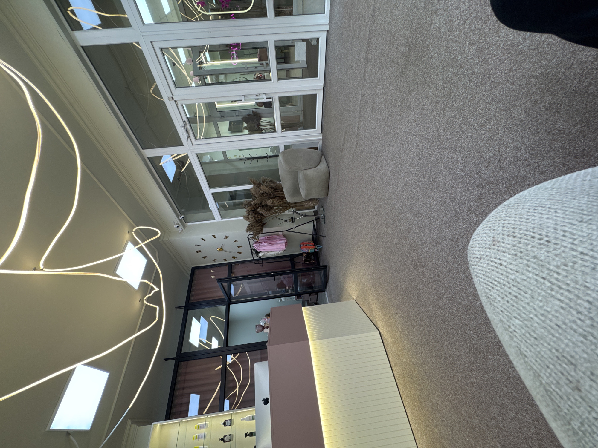 The image depicts the interior of a commercial establishment, likely a salon or reception area, in Chernivtsi, Ukraine. The space is characterized by a modern design with large glass doors and windows, a reception desk with a soft pink counter and a ribbed cream-colored base, and overhead lighting fixtures that include recessed panels and decorative illuminated tubes. The floor is covered in a textured gray carpet.
On the left, a tall glass display shelf holds various cosmetic or beauty products. A computer monitor and Apple logo are visible on the reception desk. To the right of the desk, a dark metal clothing rack displays several pink garments, and a large decorative arrangement of dried grasses stands nearby. A unique, rounded armchair is positioned to the right of the clothing rack, facing the glass doors. A clock with Roman numerals is mounted on the wall behind the rack. The glass doors reveal glimpses of an interior space, possibly an office or additional seating area. The overall ambiance is clean, contemporary, and well-lit.