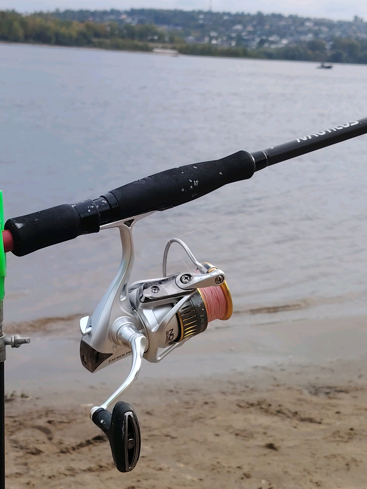The image shows a close-up of a fishing rod and reel resting on a sandy bank beside a body of water. The rod is black with white speckles on the handle and has the brand name "NAUTILUS" visible. Attached to the rod is a silver spinning reel with pink fishing line wound around the spool. The reel has a silver handle with a black grip. A green fishing rod holder is partially visible to the left of the rod. In the background, the water stretches to a distant shoreline lined with trees and buildings, suggesting an urban or suburban setting. The overall scene is consistent with a fishing location near a town or city. The location is identified as Noviy, Russia.