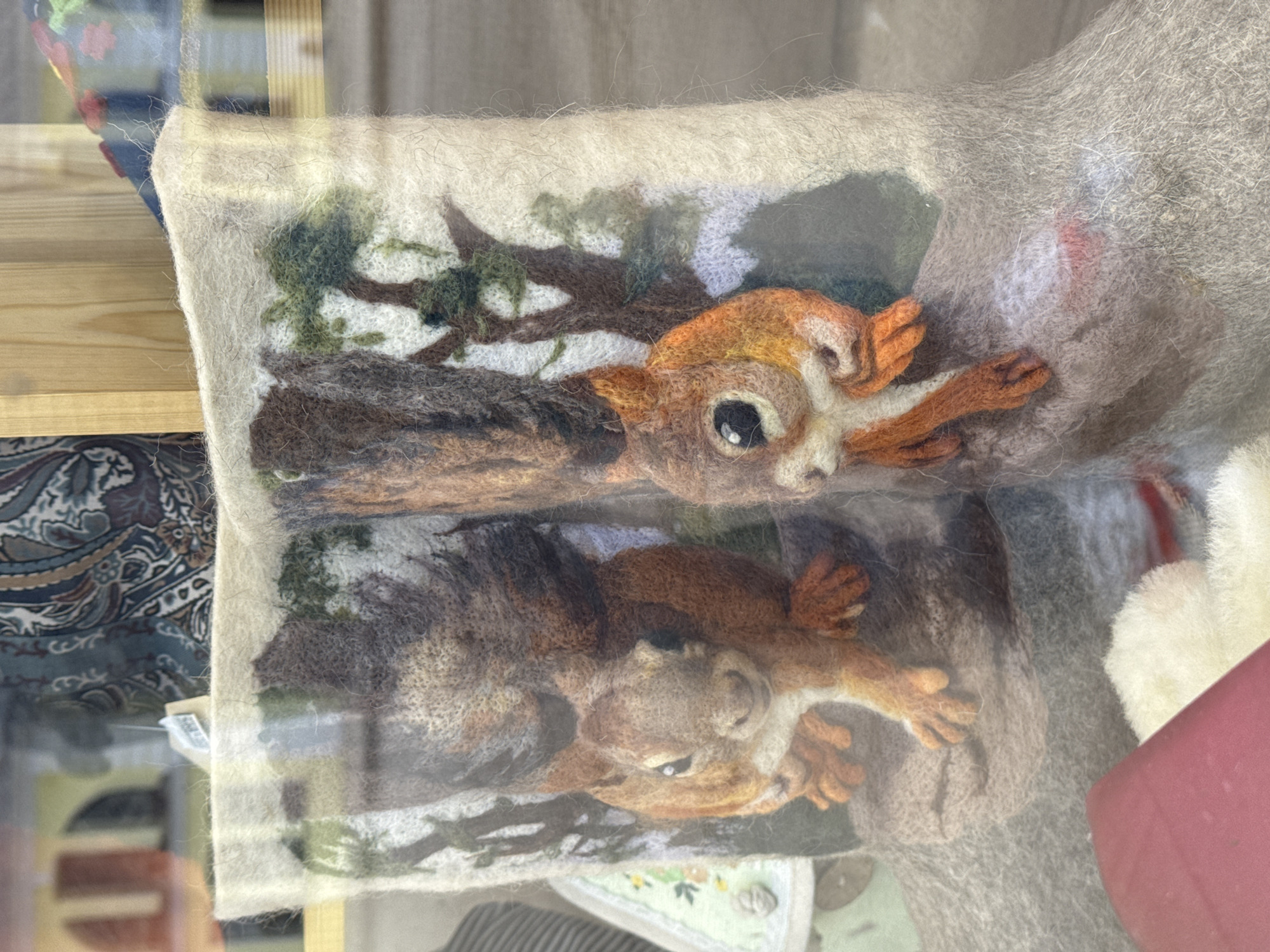 The image shows a felted wool piece depicting two squirrels on a tree branch, displayed behind a glass window. The window reflects objects from the surrounding environment, including a patterned fabric and a red container. The squirrels are rendered in shades of brown, orange, and grey, with attention to detail in their faces and paws. The background includes a suggestion of foliage in green and brown. The piece is made of felt. While the image provides visual information, it does not definitively confirm the location as Ново-Никольское, Russia, or provide specific details about people or interactions.