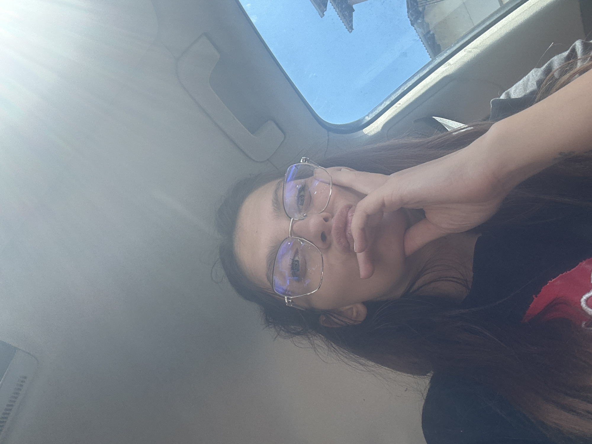 A young woman with long, dark hair and glasses is sitting in a car, looking at the camera. She has her right hand resting on her chin and her lips are pursed. Sunlight is streaming in from the right side of the car, illuminating the interior. Outside the car window, a building with a tiled roof can be seen. The location is the city of Tryavna, Bulgaria.