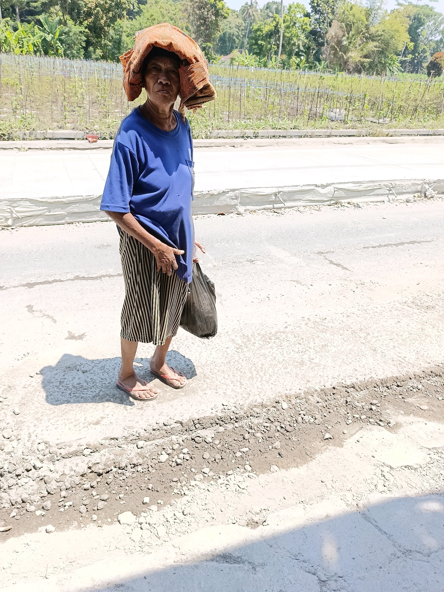 An elderly woman stands on a dirt road in Sukorini, Indonesia, on a sunny day. She is wearing a blue t-shirt and a striped skirt, with a patterned cloth wrapped around her head. In her right hand, she carries a dark plastic bag. The road is under construction, with loose gravel and some concrete visible. In the background, there are trees and a field with rows of small plants. The lighting suggests it is midday, casting a distinct shadow from the woman onto the road.