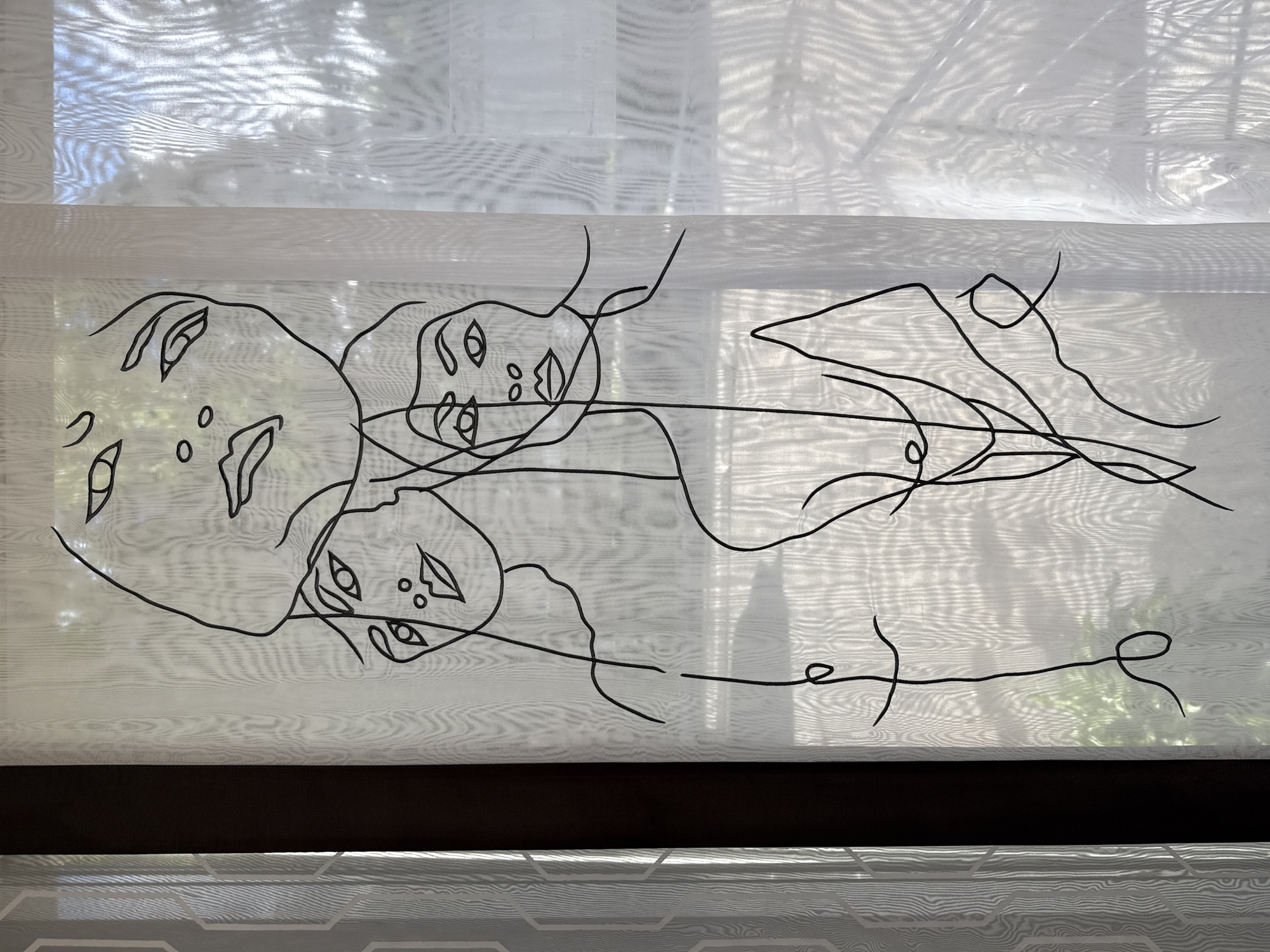 The image shows a sheer white curtain with a black abstract line drawing of three faces. The faces are superimposed, with the largest face at the top, looking downwards, and two smaller faces below it, looking forward. The drawing is continuous, with lines flowing from one face to another. The curtain is illuminated by natural light, which creates a diffused effect and casts shadows on the background. The background consists of a dark vertical element on the left and a lighter, textured surface on the right, possibly another window or wall. The overall impression is minimalist and artistic. The location is unknown, Unknown.