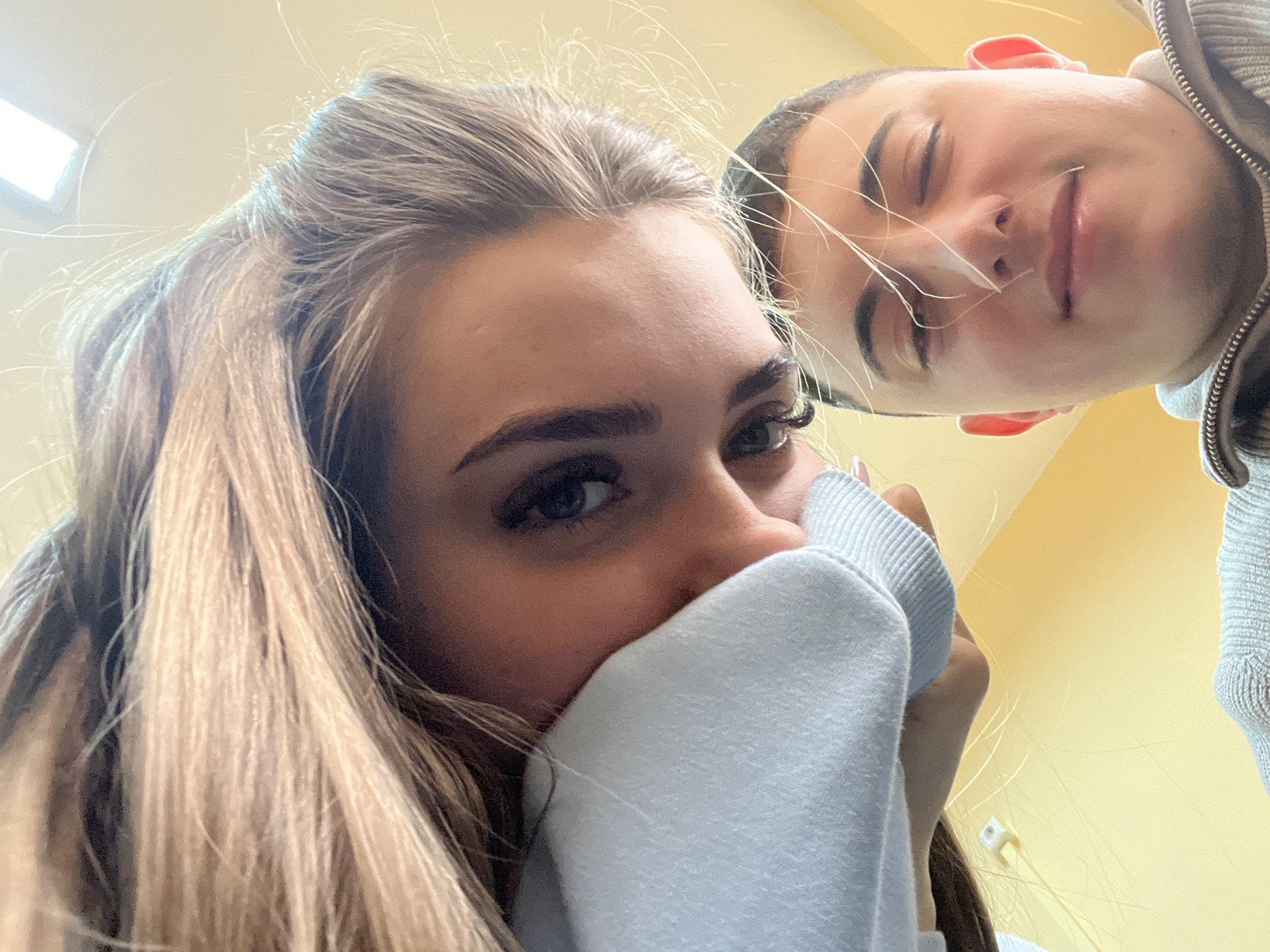 Two people are in close proximity, appearing to take a selfie. A woman with blonde hair is in the foreground, partially obscuring her face with a light-colored garment. Her eye is visible and appears blue. A man with short brown hair is slightly behind and to the right of the woman. He has his eyes closed and a slight smile. The background is a plain yellow color. There is no evidence to suggest the image was taken in Nova Zagora, Bulgaria.