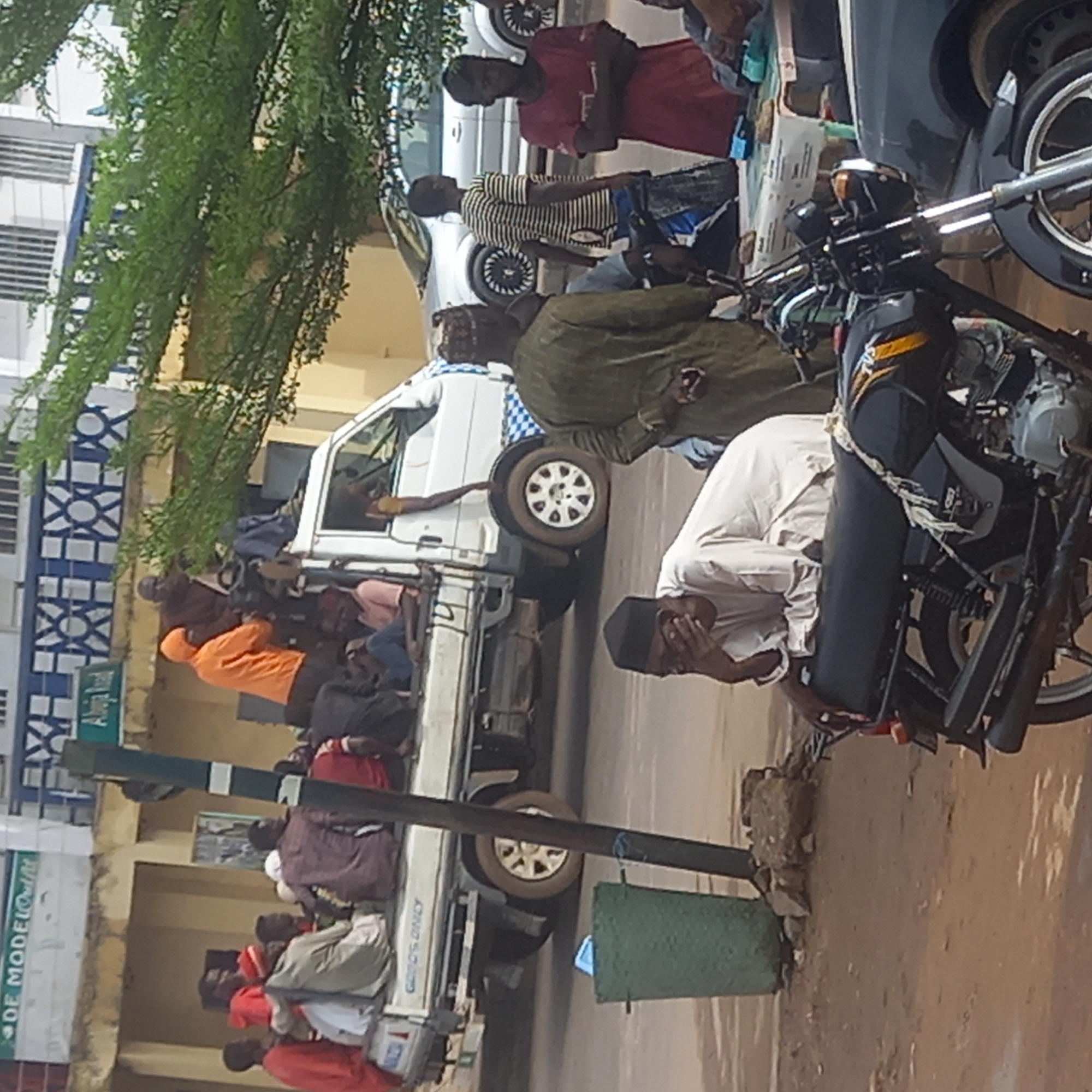A daytime, medium shot, photographed horizontally, depicts a street scene in Dadi Riba, Nigeria. A group of people is riding in the back of a white pick-up truck. Many individuals are dressed in brightly colored clothing. A man in a white robe and a black hat is near a motorcycle. Several other vehicles and people are present in the background. The light quality appears to be bright and somewhat harsh. A tree is visible in the upper left of the frame.