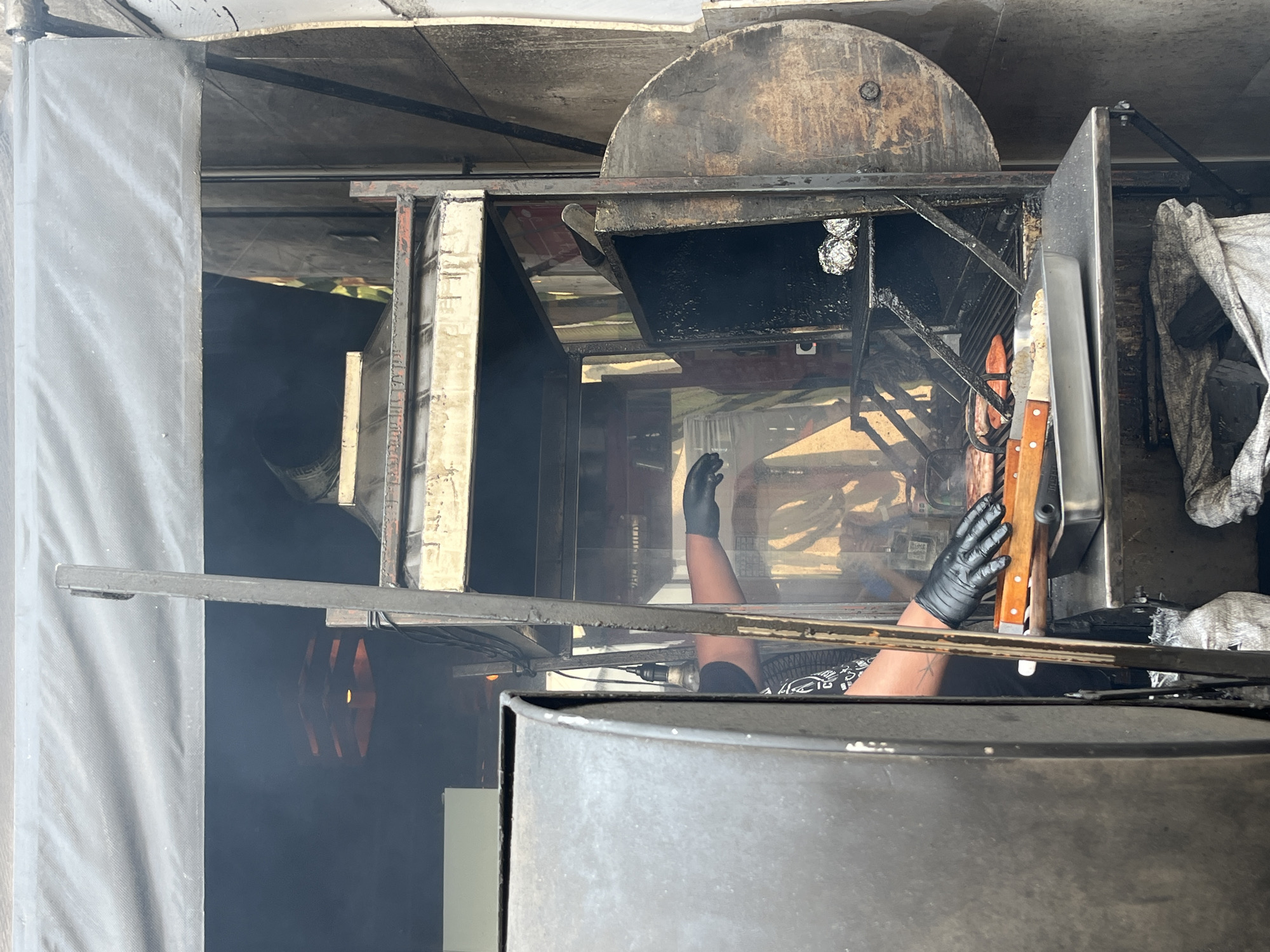The image depicts a person wearing black gloves inside what appears to be a large metal barbecue smoker or grill. The person's arms are visible, reaching into the smoker. Several cooking tools, including knives with wooden handles, are visible, resting on a metal surface. The interior of the smoker is dark and greasy, with some aluminum foil balls visible on a rack. There is smoke present, indicating the smoker is in use. The exterior of the smoker appears weathered and used. A grey cloth or tarp is partially visible on the right. The environment appears to be an outdoor cooking setup, likely a food stall or catering operation. Details to suggest the location is San Francisco Totimehuacán, Mexico are not discernable in the image.