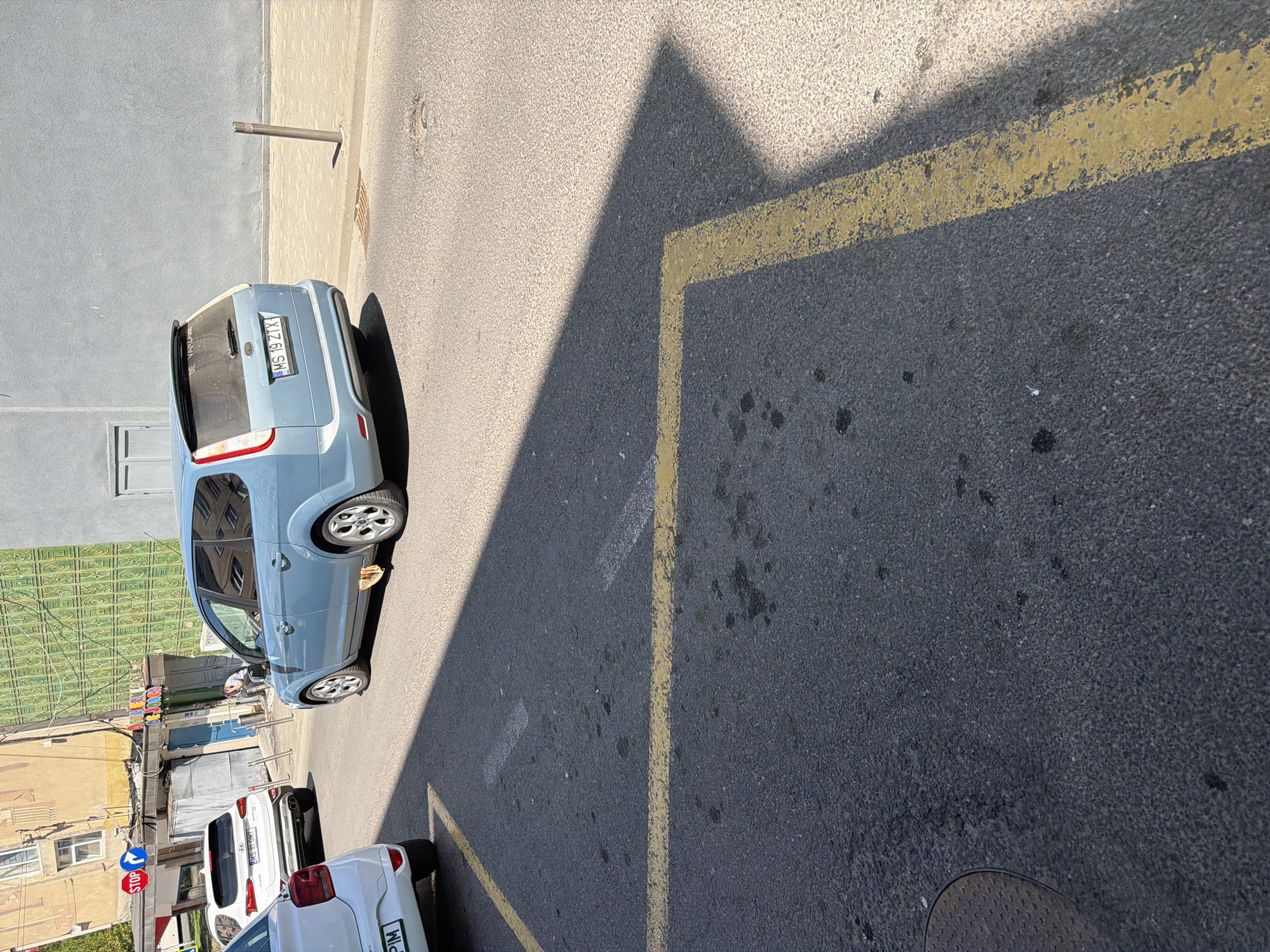 The image shows a street scene in Târgu Mureș, Romania. A light blue hatchback car with the license plate "MS 19 ZIX" is parked beside a sidewalk. There is a yellow parking line and triangle shadow on the road. There are two additional cars further down the road. There is a building on the left side. In the distance are street signs and an intersection.