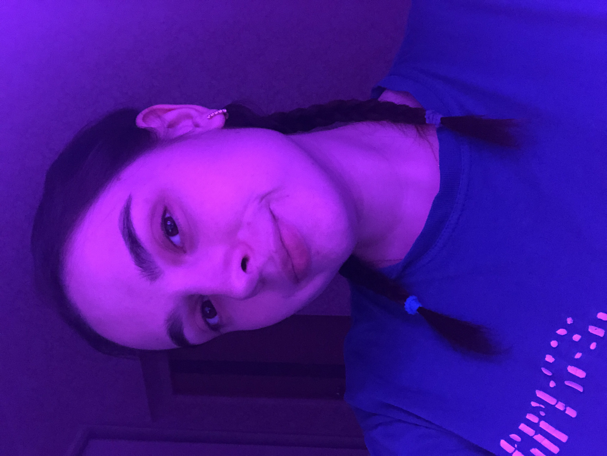 A female subject is depicted in a close-up, chest-up portrait within an indoor environment illuminated by a strong purple/magenta light source. The subject, a young adult female with fair skin, has dark hair styled in two braids, visible over each shoulder, with light blue hair ties near the ends of the braids. She has dark eyebrows and dark eyes, and a slight, closed-lip smile. A crescent-shaped earring with multiple reflective elements is visible on her right earlobe. She is wearing a blue crew-neck t-shirt featuring a graphic design on the front composed of irregular pink/magenta shapes and dots that appear fluorescent or UV-reactive under the purple lighting. The subject gazes directly at the camera. The background is indistinct, showing a dark wall and a vertical structural element, possibly a door frame or paneling, on the left side of the frame. The specific location in the city of Noviy, Russia, cannot be determined from the visual information provided in the image.