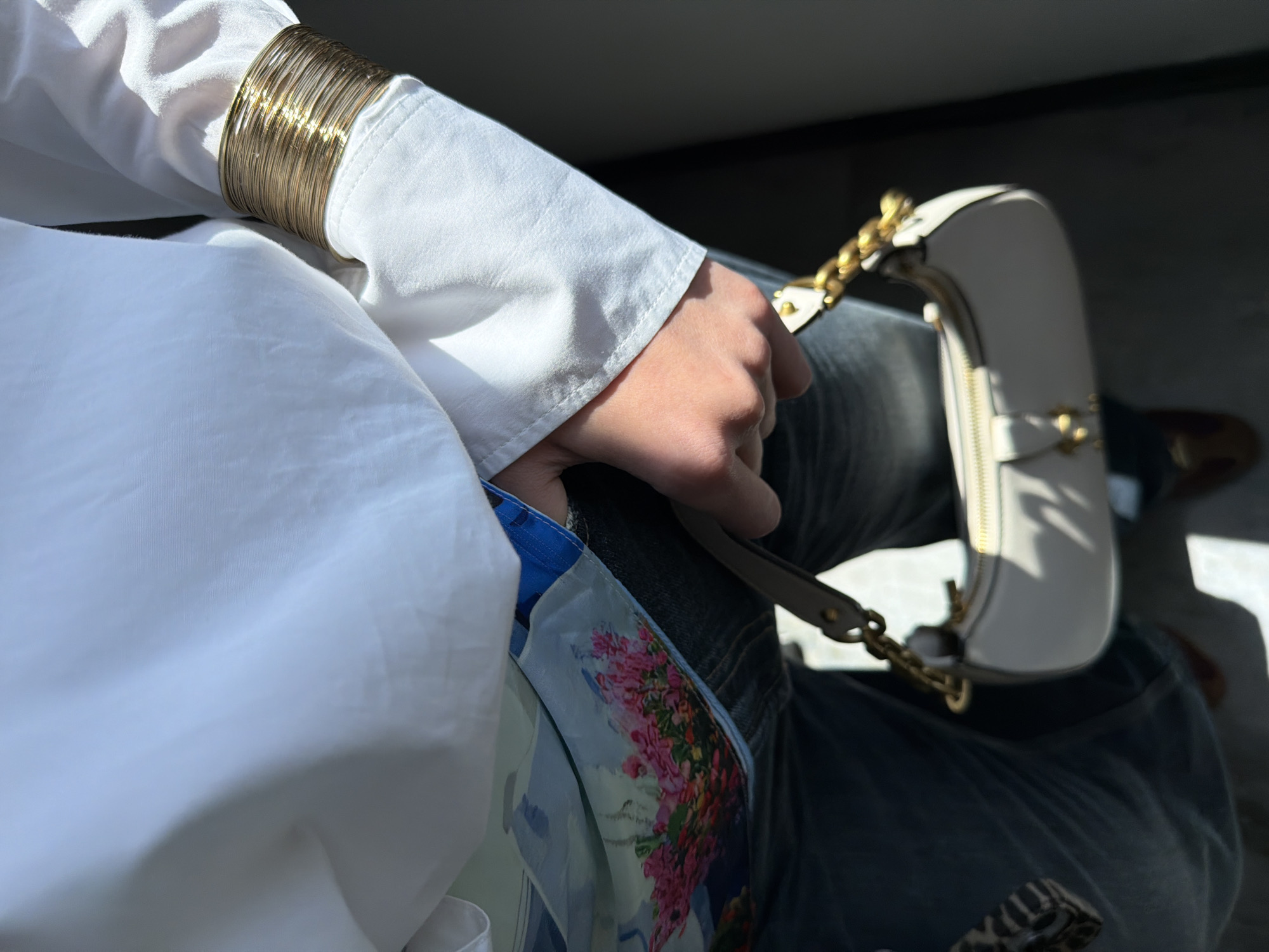 The image captures a close-up, high-angle view of a person seated, focusing primarily on their arm, hand, and lap. The subject is wearing a white long-sleeved shirt or blouse, and blue denim jeans. On their right wrist, a stack of multiple gold bangle bracelets is prominently displayed, glinting in the sunlight. Their hand lightly grips a small, white handbag with a gold chain strap and a gold zipper detail.
A colorful scarf, featuring a vibrant floral pattern with shades of pink, red, and blue, is partially visible tucked into the lap area, contrasting with the crisp white shirt. The lighting is characterized by strong, direct sunlight streaming in from the left, creating bright highlights on the white fabric and gold accessories, and casting deep shadows in the background. This high-contrast lighting suggests it is daytime. The background is dark and indistinct, hinting at an indoor setting or a vehicle interior. The person appears to be resting or traveling. No text is visible in the image. The scene is set in Yerevan, Armenia.
