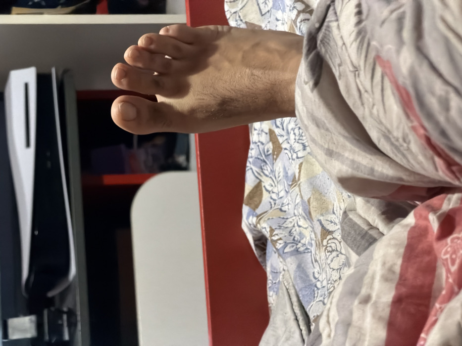 The image captures an indoor scene in Opanets, Bulgaria, viewed from a low perspective, likely from a bed. In the immediate foreground, patterned bed linens are visible, featuring grey, white, and red stripes, as well as a blue, brown, and white floral design. A bare human foot, likely male due to visible hair on the instep, extends upwards from the blankets. The foot exhibits five digits with visible nails. Directly behind the foot, a flat red surface is present, possibly part of a bed frame or furniture. In the background, a white shelving unit houses a horizontally positioned white and black PlayStation 5 console. To the left of the console, several dark electronic devices are stacked. A red compartment is also discernible within the shelving unit to the right.
