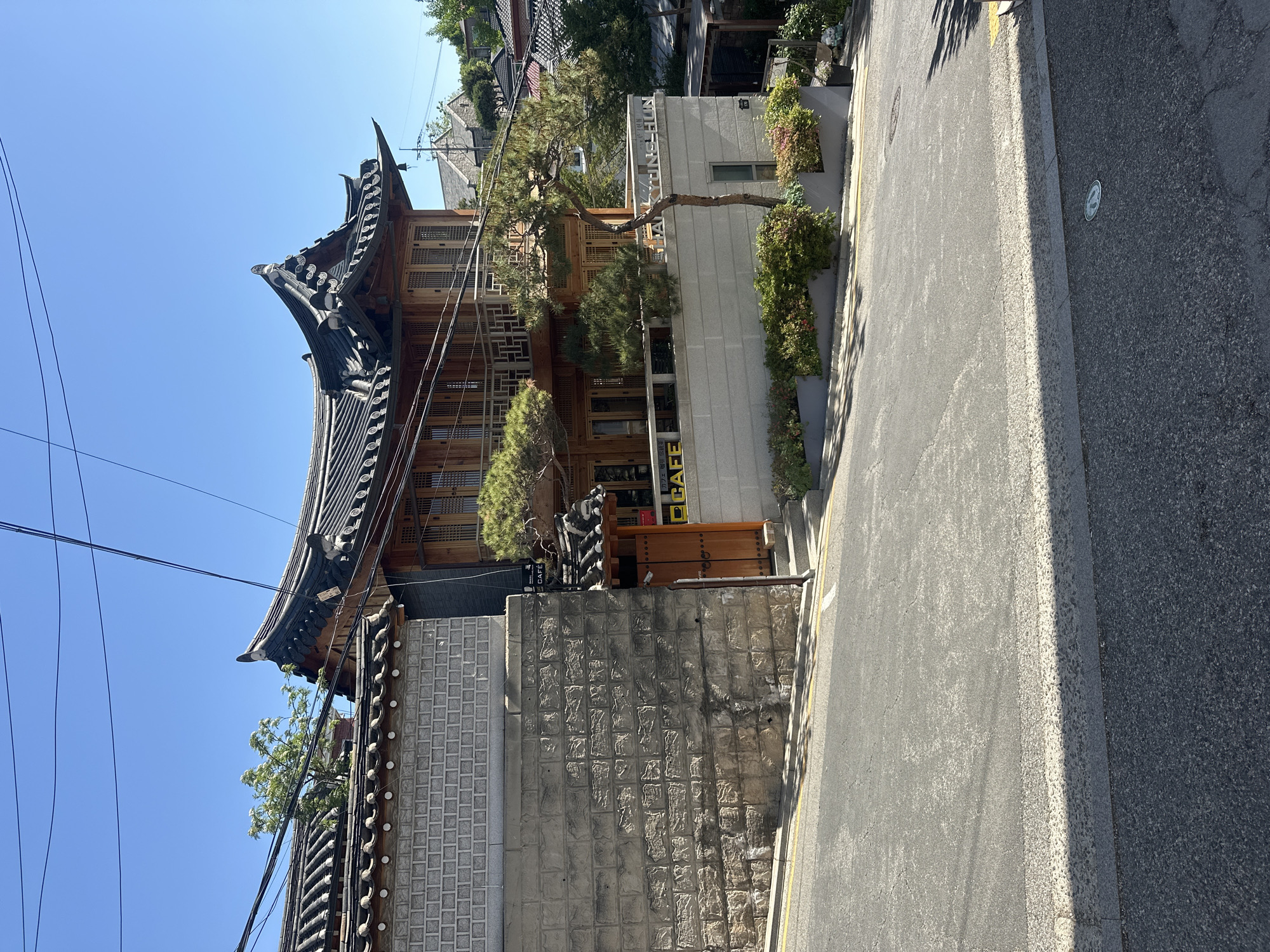 The image shows a traditional Korean building in Seoul, South Korea. The building features a dark tiled roof with curved eaves, wooden frame construction, and traditional Korean windows. The building appears to be a cafe, as indicated by a sign visible. The cafe is located adjacent to a gray brick wall and a paved street. Other buildings are seen in the background. The sky is blue. Overhead cables are strung above.