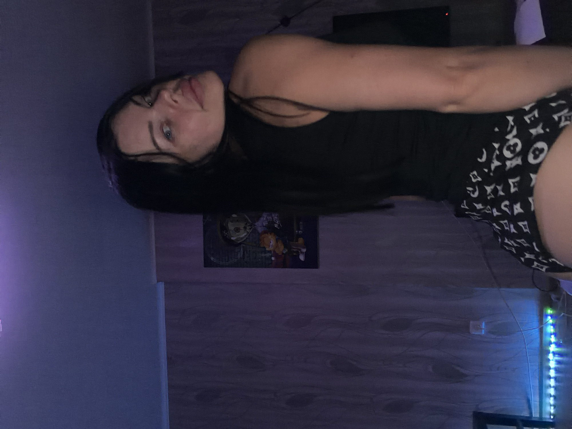 A young woman with long, dark hair and blue eyes is shown in a dimly lit room. She is wearing a black tank top and patterned shorts with a white logo. Her head is tilted back, and she appears to be looking upwards. The room has wood-paneled walls and is decorated with a framed picture on the wall. There are also LED lights visible, casting a colorful glow. The time of day appears to be evening or night due to the lighting conditions. The overall mood is relaxed and casual, suggesting the woman is at home. There are no identifiable landmarks to suggest the specific location within Ufa, Russia, beyond the provided context.