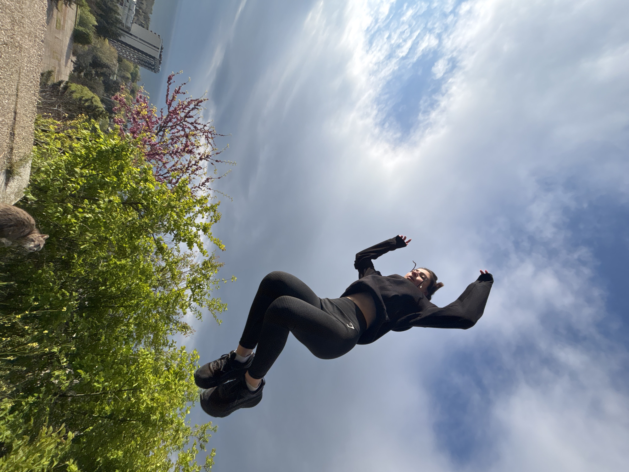 An outdoor, low-angle photograph captures a person suspended in mid-air against a bright, partly cloudy sky. The image, originally rotated, shows the central subject wearing black athletic attire: a long-sleeved crop top, black leggings with a white "GYMSHARK" logo visible on the left thigh, and black sneakers. Their body is dynamically posed, slightly bent with arms raised, giving the impression of a playful jump or a moment of weightlessness.
The left side of the frame is rich with natural elements, featuring vibrant green foliage from trees or bushes, and a distinct tree with pink and purple blossoms. A textured grey wall or structure runs along the far left edge of the scene. Partially visible in the top-left corner, peeking from behind the greenery, is the head of a grey cat. In the distant background, beyond the trees, an urban landscape unfolds with a large, multi-storied grey building and a calm body of water (possibly a lake or sea) meeting the horizon. The sky, which fills the majority of the background, displays a mix of soft, wispy white clouds against patches of clear blue, indicating a bright, pleasant daytime. The overall impression is one of dynamic movement set against a blend of urban and natural scenery.