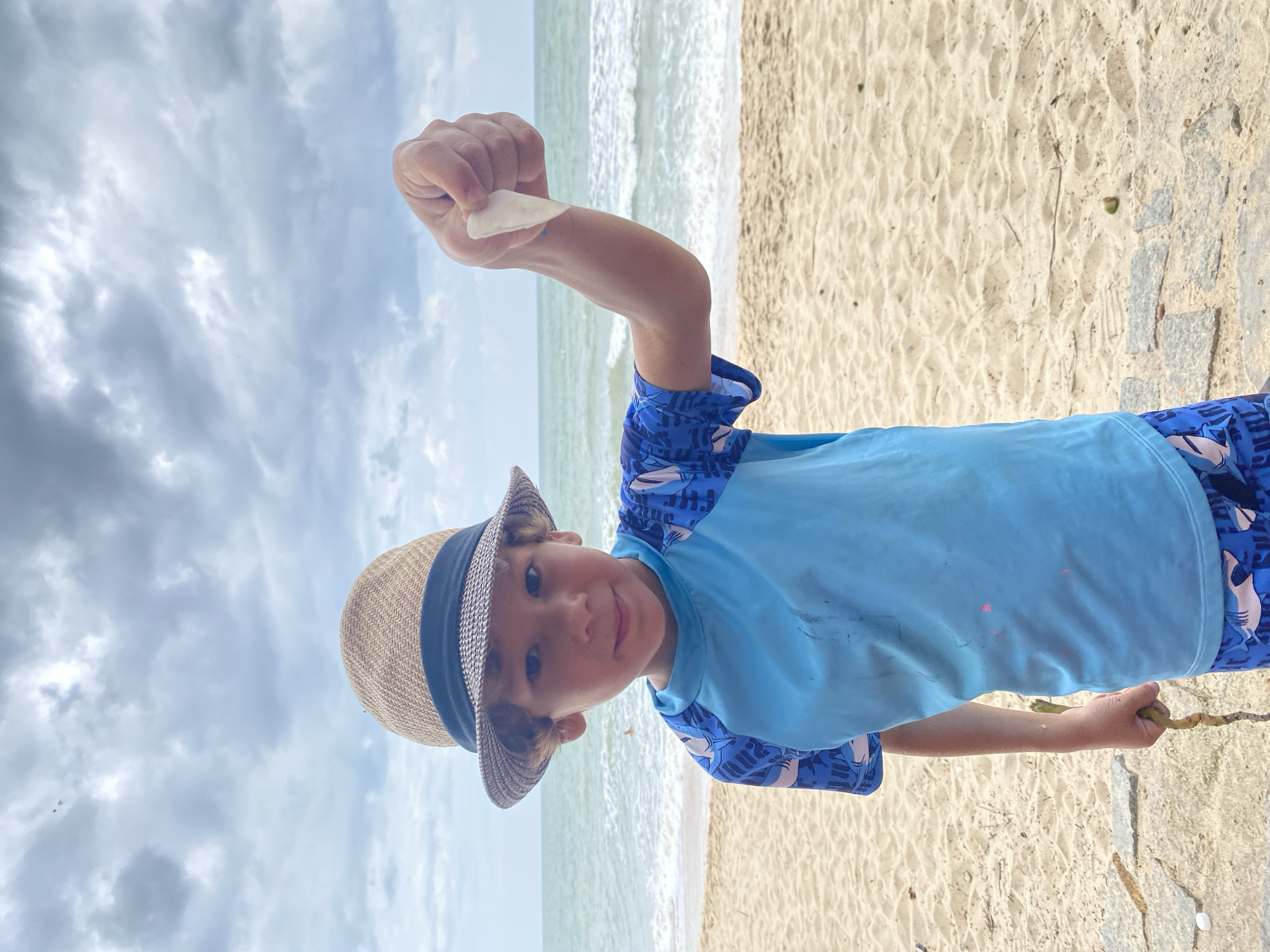 A young boy wearing a straw hat and a blue rash guard with shark patterns stands on a sandy beach in Mũi Né, Vietnam, holding a piece of white shell in his raised right hand. The boy is looking towards the camera with a slight smile. Behind him, the ocean with gentle waves stretches towards the horizon under a cloudy sky. The sand is light brown and textured, with a few scattered stones and debris. The boy's left hand holds a piece of driftwood.
