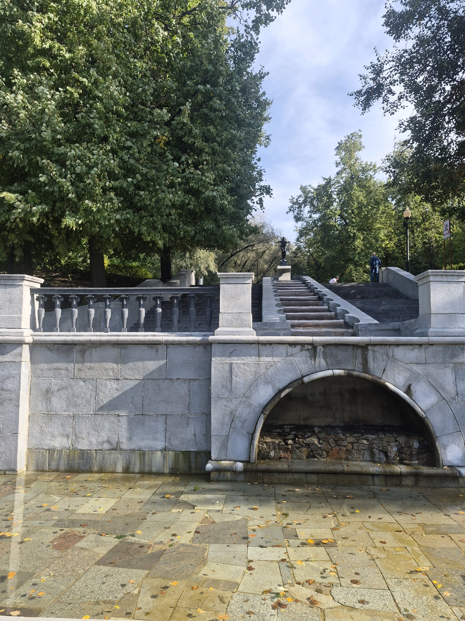 This image depicts a wide stone staircase leading upwards, flanked by monumental stone structures and a balustrade. The staircase is situated in a park-like setting with mature trees and a bright, partly cloudy sky. In the foreground, a shallow pool of water reflects the light and fallen leaves, creating a mosaic of colors on the stone paving. The architecture suggests a formal public space, possibly a memorial or a decorative element within a larger park. The presence of a few individuals on the stairs and the overall ambiance indicate it is daytime and the weather is pleasant. The stone balustrade, ornate pillars, and the semicircular archway beneath the stairs point to a neoclassical or Beaux-Arts design. The scene is observed from a low angle, emphasizing the grandeur of the staircase and the surrounding stonework. The overall impression is one of serene public beauty, with a touch of autumn foliage. This scene is located at the Alexander Garden in Moscow, Russia.