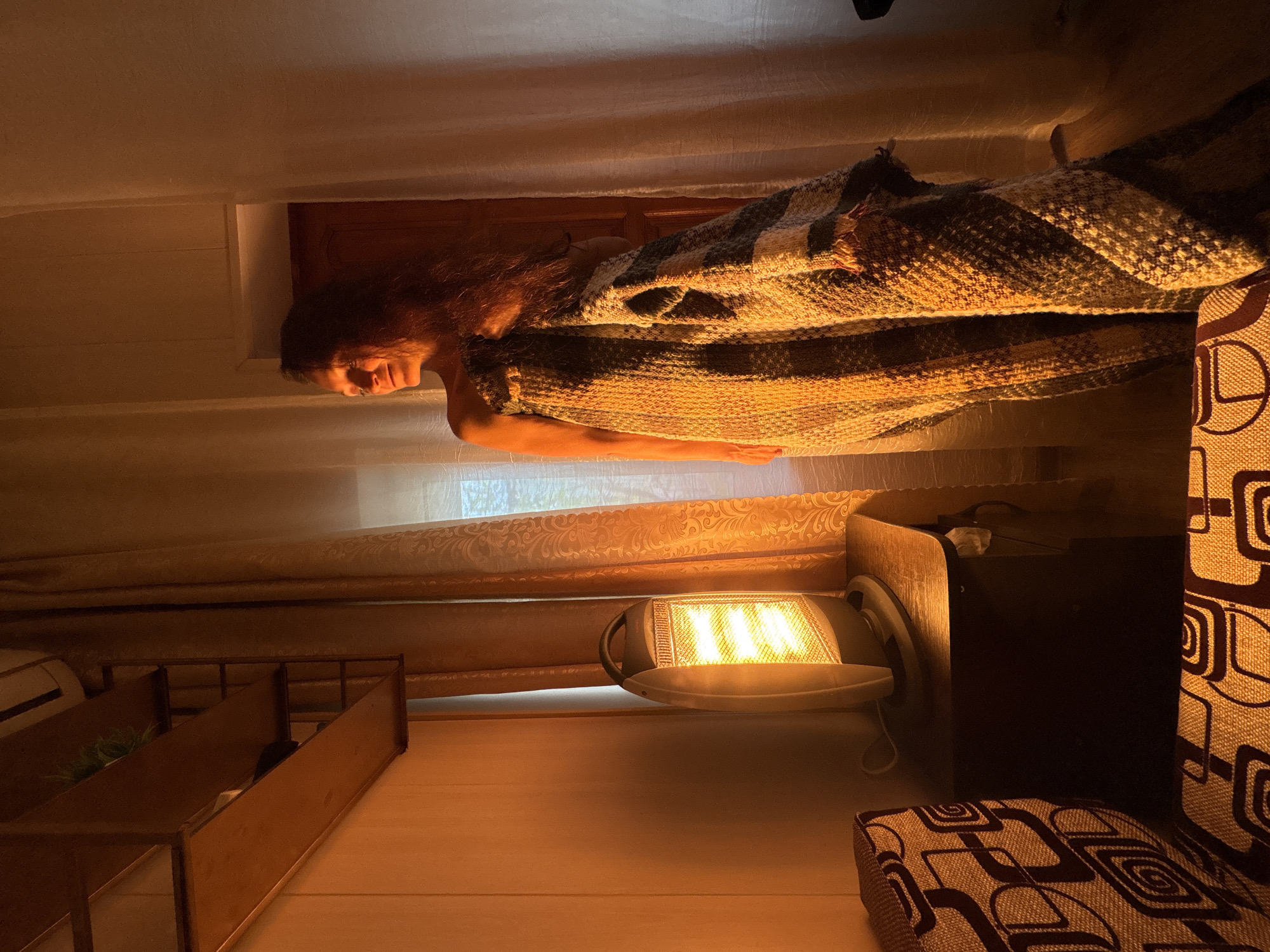 A woman lies in bed, covered with a blanket, in a room illuminated by an electric heater. The room contains furniture, including a bedside table and a patterned chair. The location may be Gelendzhik, Russia.