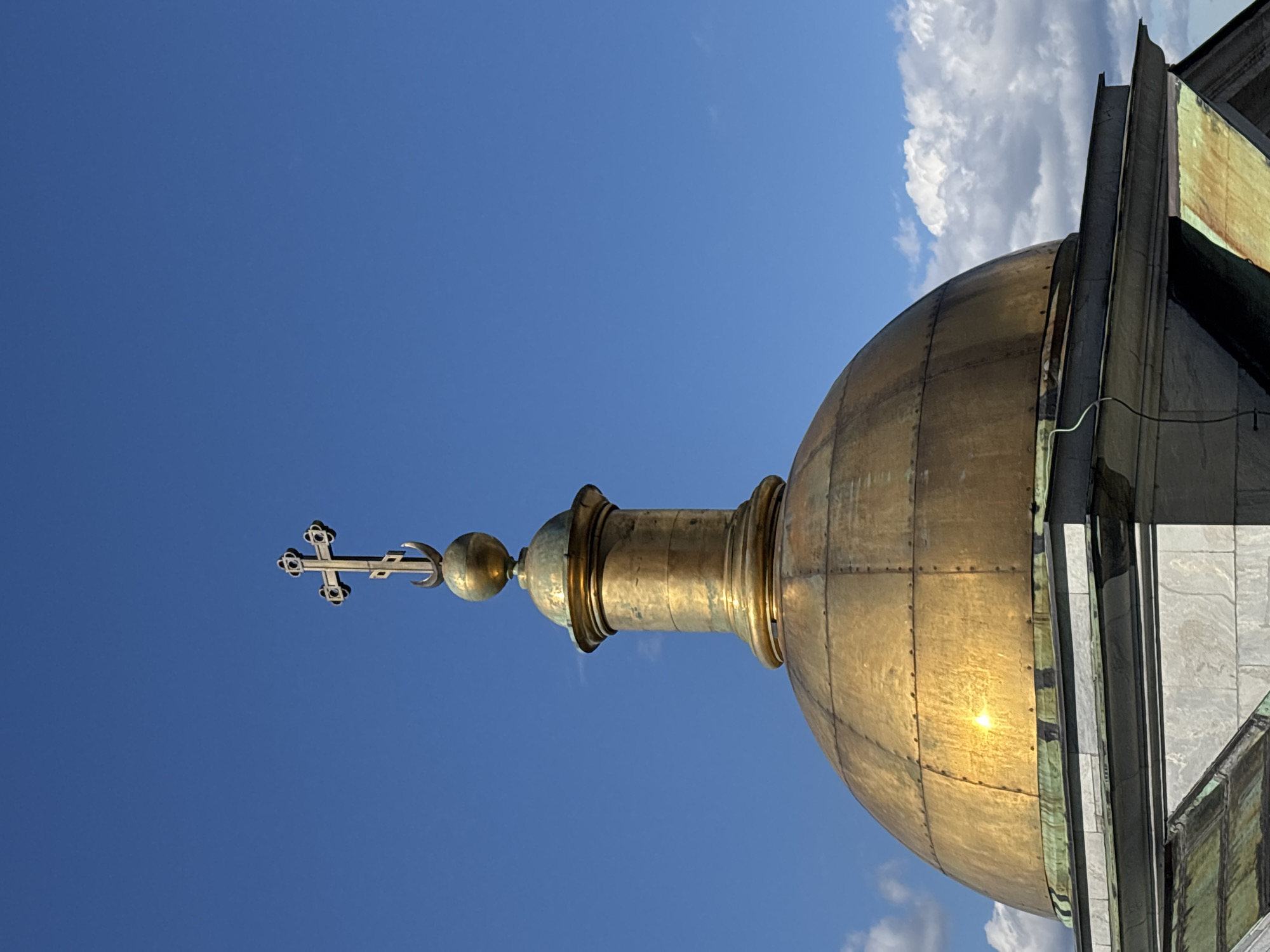 The image displays the top portion of a golden dome and a cross, set against a bright blue sky with scattered clouds. The dome is made of metal panels and is reflecting the sunlight. A cross with intricate decorative elements is positioned above a spherical finial on a slender spire, which is in turn attached to the dome. The architecture appears to be of a religious building, possibly a church or cathedral. The sun's glare on the dome suggests it is daytime, likely midday, given the bright, clear sky. The scene evokes a sense of grandeur and spiritual significance. There is no visible text in the image.