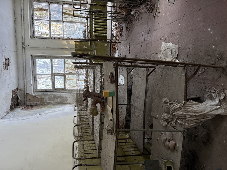 An abandoned room features rows of dilapidated metal bunk beds, coated in dust and debris. Two large windows illuminate the space, showcasing cracked walls and peeling paint. A doll rests on one bed, adding an unsettling element. Discarded materials and remnants of past occupants litter the floor. The room exudes a sense of decay and neglect.