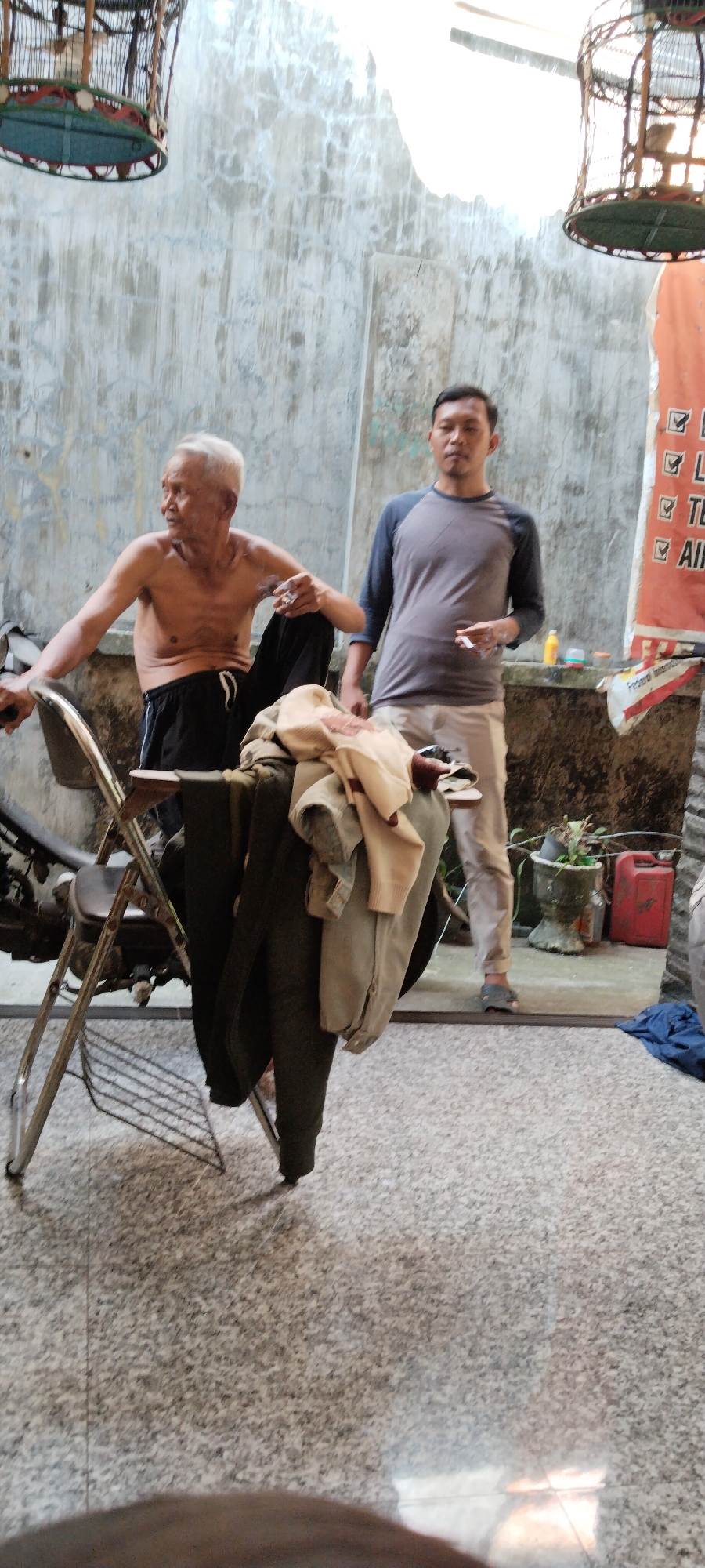 In Peganjaran, Indonesia, two men are observed near a concrete wall. An elderly man, shirtless and with white hair, is seated on a metal chair, holding a cigarette and appearing to look to his left. He is wearing dark pants. A younger man, wearing a grey long-sleeved shirt and light-colored pants, stands to the right of the elderly man, also holding a cigarette. In the foreground, a pile of clothing, including dark trousers and lighter-colored garments, is draped over a chair. Behind the men, a birdcage hangs from above. The ground is a speckled terrazzo-like surface. To the far right, a red jerrycan and a potted plant are visible near the wall. A partially visible orange banner with some text and checkmarks is also present.