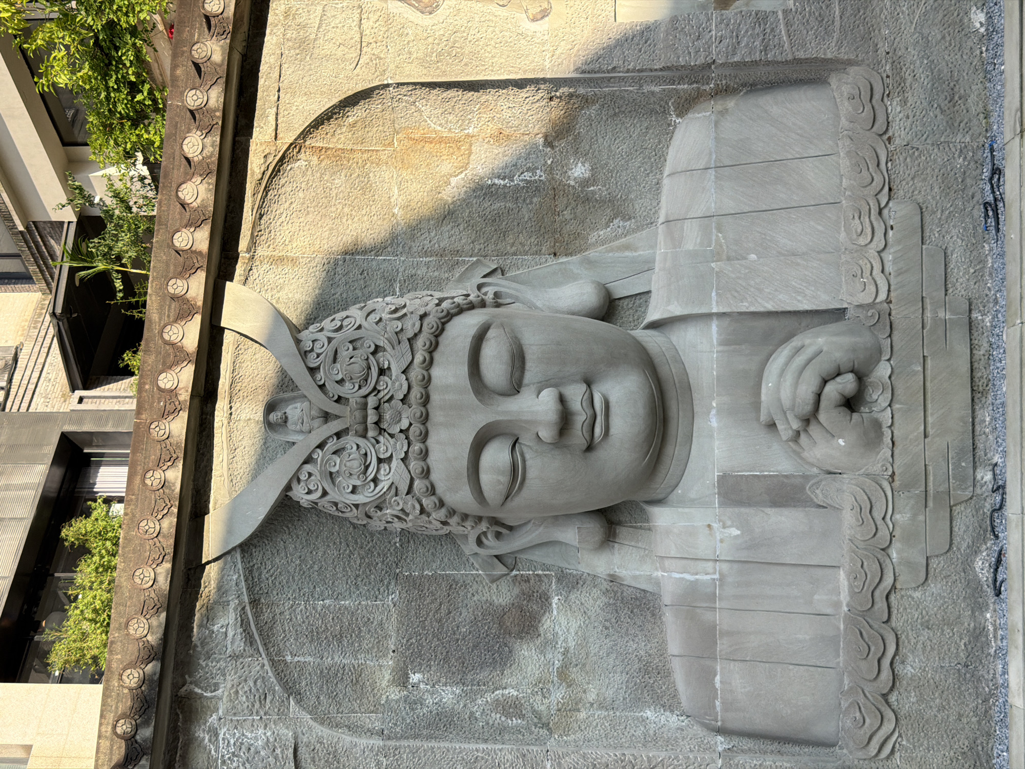 The image depicts a large, intricately carved stone relief of a Buddha on a textured stone wall. The Buddha is depicted with closed eyes, a serene expression, and wearing an ornate headdress. Its hands are clasped in front of its chest. The relief is set within an arched recess, and above it, a decorative stone frieze runs horizontally. Behind the frieze, a building with windows and lush green foliage is visible. The sun casts strong shadows across the relief and the wall, indicating it is likely daytime. This relief is located at the Guangming Temple in Nan'an District, China.