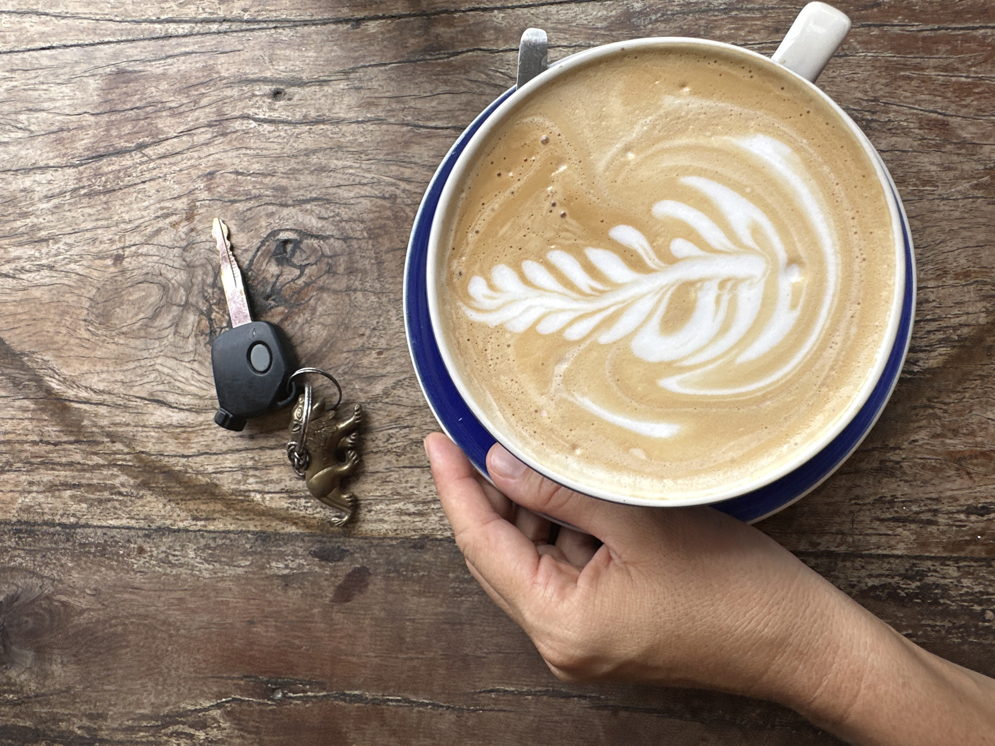 A close-up, overhead shot shows a person's left hand holding a white coffee cup with a blue rim, filled with latte art, on a weathered wooden table. To the upper right of the coffee cup, a car key with a black fob and a small brass lion keychain are visible. The background is a rustic wooden surface with distinct grain patterns. The scene appears to be in a cafe or outdoor seating area on Koh Phangan, Thailand, given the context of the location.