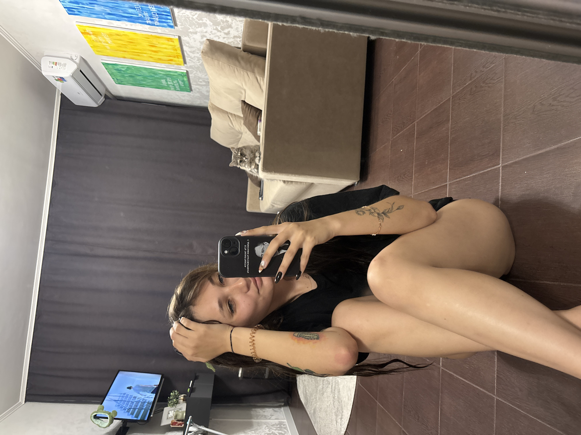 A young woman with long brown hair is sitting on a dark wood floor in what appears to be a living room. She is wearing a black t-shirt and has several tattoos on her arms and legs. She is holding her phone in her right hand and taking a selfie. In the background, a beige sofa is visible with a cat sitting on it. The room also contains a television mounted on the wall, an air conditioner, and several colorful paintings on the wall. The overall impression is that of a casual indoor setting. No specific details indicate the location is in Novokuznetsk, Russia.