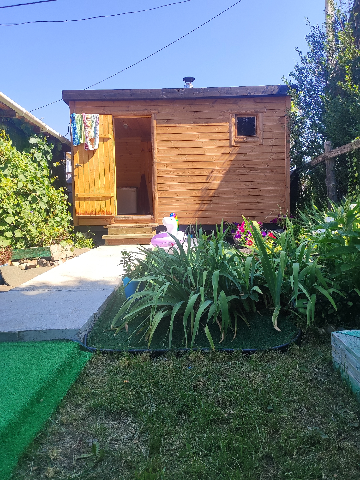 The image depicts a small wooden structure, possibly a shed or sauna, in a backyard setting under a clear blue sky. The structure is made of horizontal wooden planks and has an open doorway with steps leading up to it. Three colorful towels are hanging on the left side of the doorway. A small window is visible on the right side of the structure. In front of the structure, there is a patch of lush green plants and flowers, including some with pink and purple blooms. To the left of the structure, there is a concrete path leading to a garden area with green foliage. The foreground shows a lawn with artificial turf and natural grass. The scene suggests a well-maintained outdoor space. No specific details about the city of Ridder, Kazakhstan, are visible in the image.