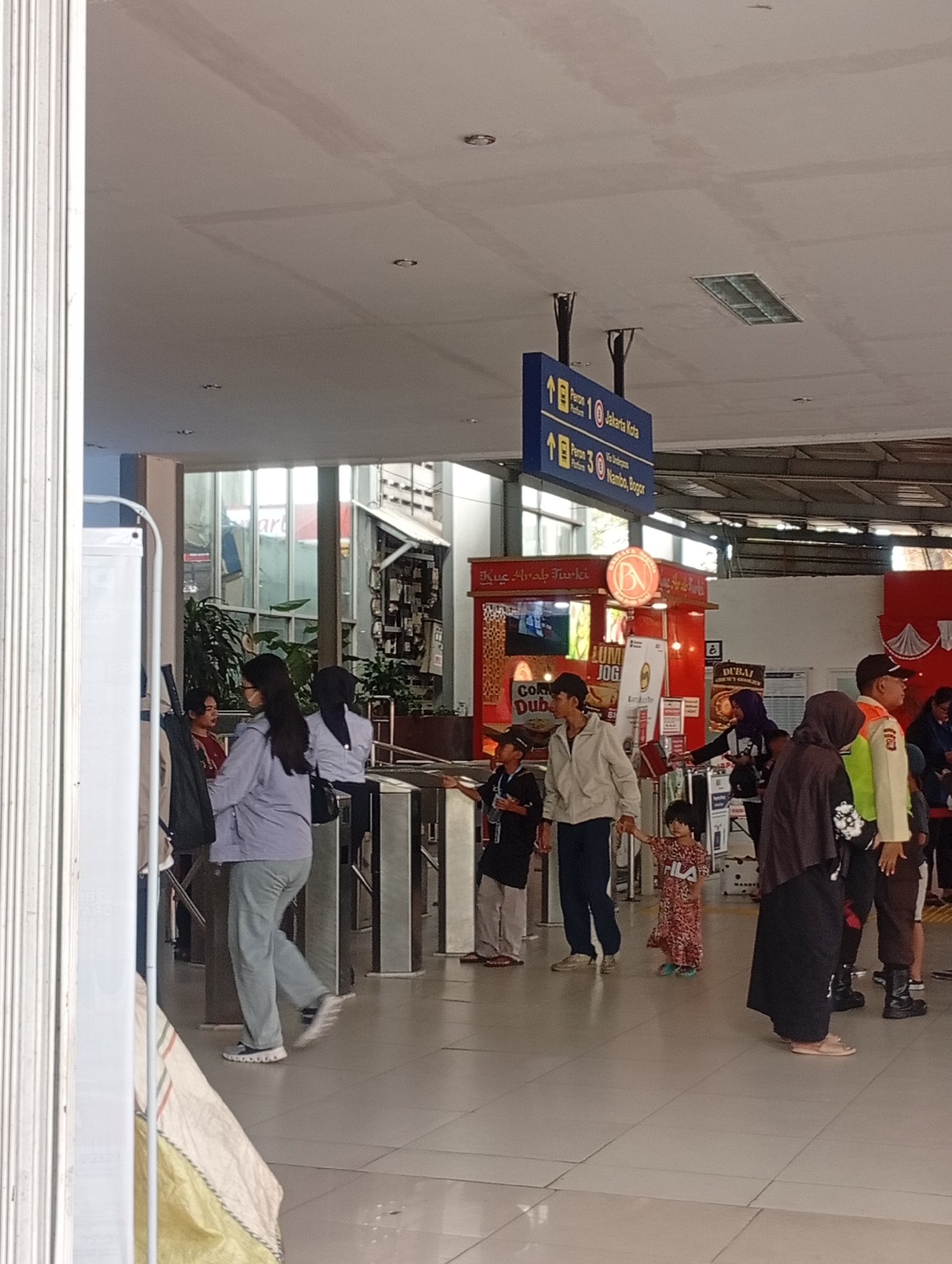 The image depicts a busy interior scene, likely at a transportation hub in Depok, Indonesia. In the foreground, a group of people are seen navigating through turnstiles. A woman in a light purple jacket and grey pants is walking towards the right, with a black bag slung over her shoulder. To her left, another woman in a similar jacket and grey pants is partially visible. A young child in a patterned dress is being held by the hand by a man wearing a light-colored jacket and dark pants. Behind them, a man in a beige jacket and dark pants stands next to a young boy in a black shirt and cap.
Further back, a food stall with red and white branding, displaying text like "Kuc. Arab Turki" and "LUM JOG", is visible. Above the crowd, a blue directional sign indicates "Peron 10 Jakarta Kota" and "Peron 30 Nambo, Bogor", confirming the location as a train station or terminal. Several other individuals are seen in the background, including a person in a security uniform with reflective green and orange accents. The environment appears to be indoors, with a tiled floor and a ceiling with visible lighting fixtures. The overall atmosphere suggests a public space with people in transit.