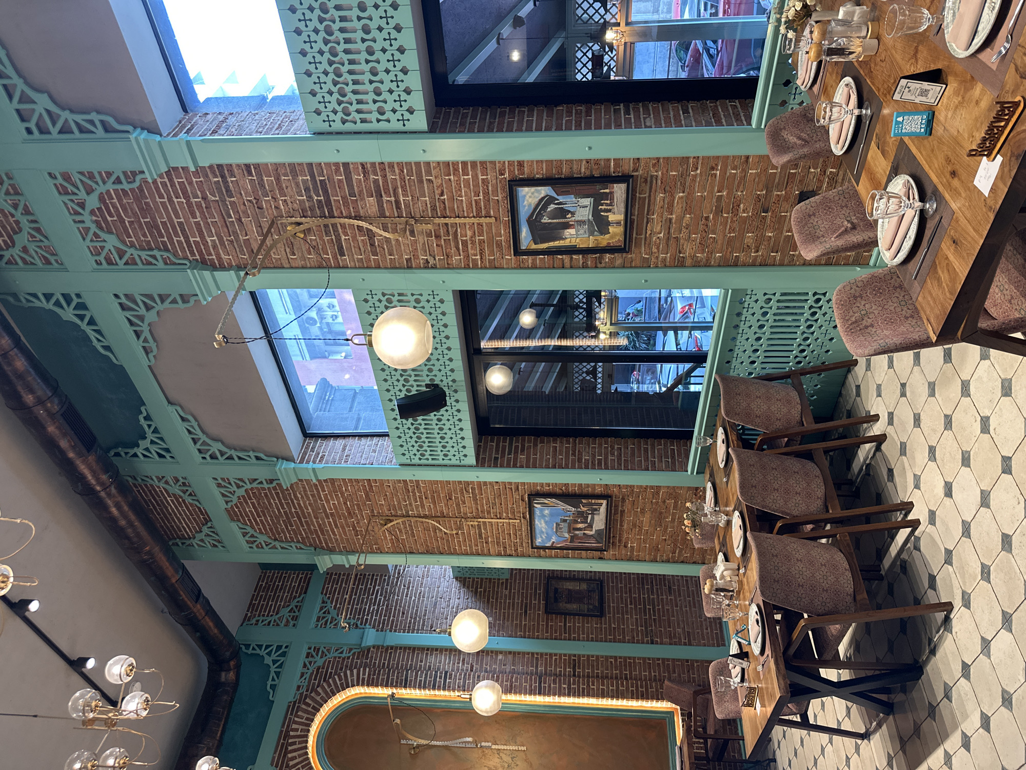 The image depicts the interior of a restaurant, likely in Batumi, Georgia, based on the architectural style. The restaurant's design is characterized by brick walls interspersed with light green, ornate, geometric-patterned structures that appear to be structural beams or decorative elements. Two framed pictures hang on the brick walls. The ceiling features multiple light fixtures, including hanging spherical lights. The floor is tiled with a geometric black-and-white pattern. Tables are set with plates, cutlery, and glassware, and chairs are upholstered in a patterned fabric. A "Reserved" sign is visible on one of the tables, indicating that the table is not available for general seating. Natural light enters through the windows. The overall atmosphere seems upscale and designed with an eye for detail.